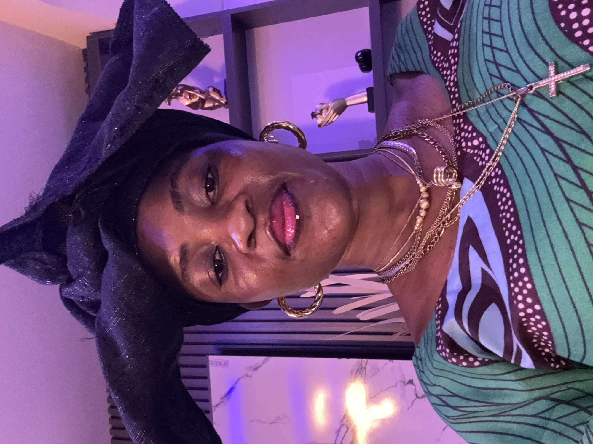 In a medium shot, a woman with dark skin and a dark headwrap is positioned centrally. She has on a green and blue patterned top adorned with multiple gold necklaces. Her eyes are directed towards the camera, and she has a subtle smile on her face, with pink lipstick and a lip piercing visible. To her left, a wall is illuminated with purple light, and to her right, shelves hold decorative items. The background appears to be an indoor setting.