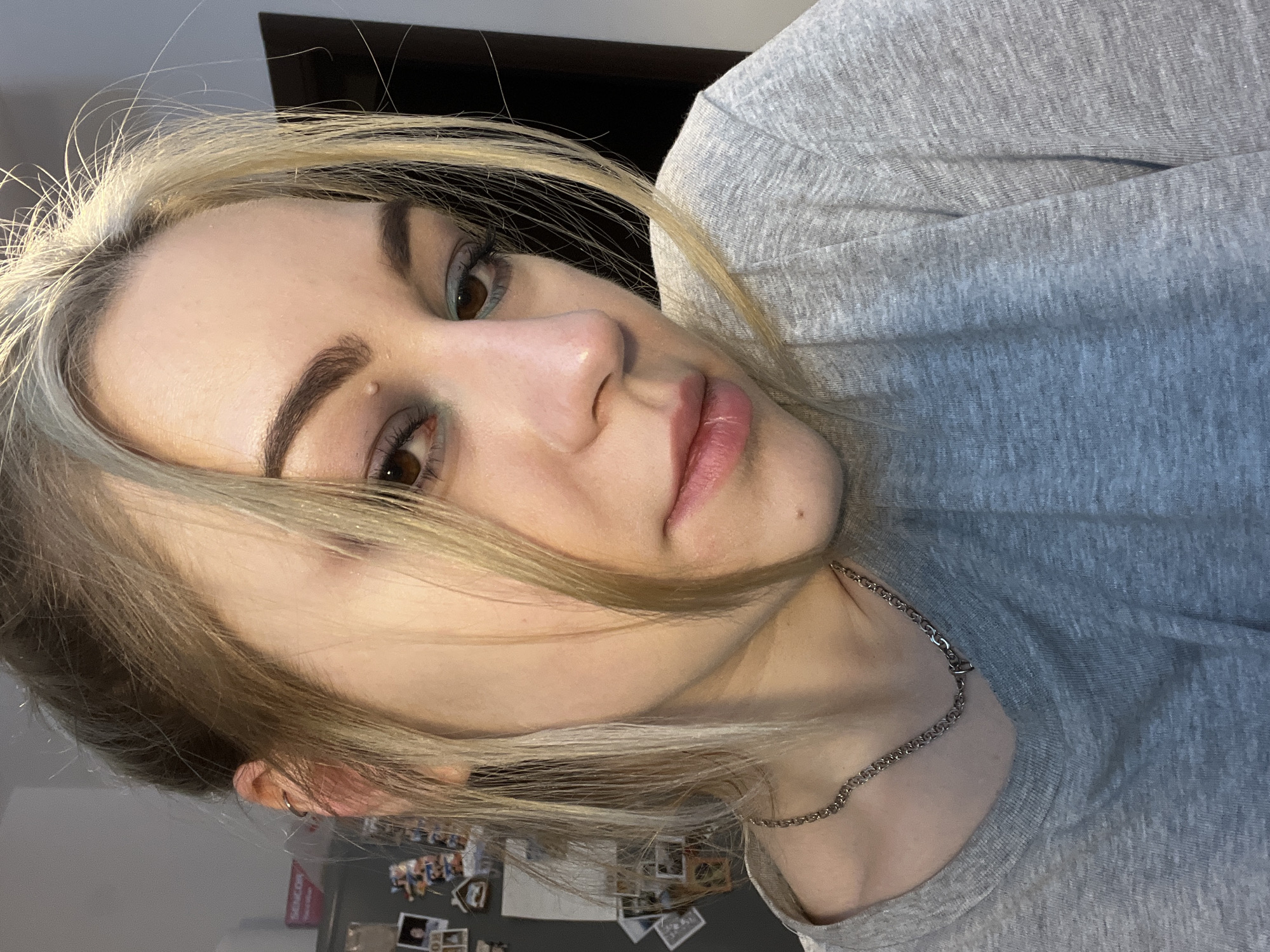A close-up, tilted, indoor portrait of a young woman with blonde hair and brown eyes. She wears a gray t-shirt and a silver chain necklace. Her makeup is visible on her eyes. A mole is present on her forehead, and another on her neck. A cluttered wall is visible in the background with pictures/postcards affixed to the wall. The location in Boreti, Montenegro is not discernible from the image.