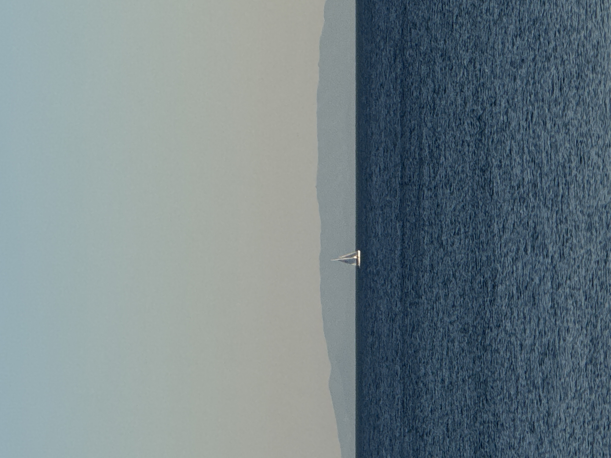 A sailboat with a white sail is pictured in the distance on the sea, with a hazy mountainous landmass in the background. The sea is a deep blue, with subtle ripples on its surface. The sky above is a pale, gradient blue, transitioning to a lighter hue near the horizon. The scene is serene and expansive. The location is the coast of Alimos, Greece.