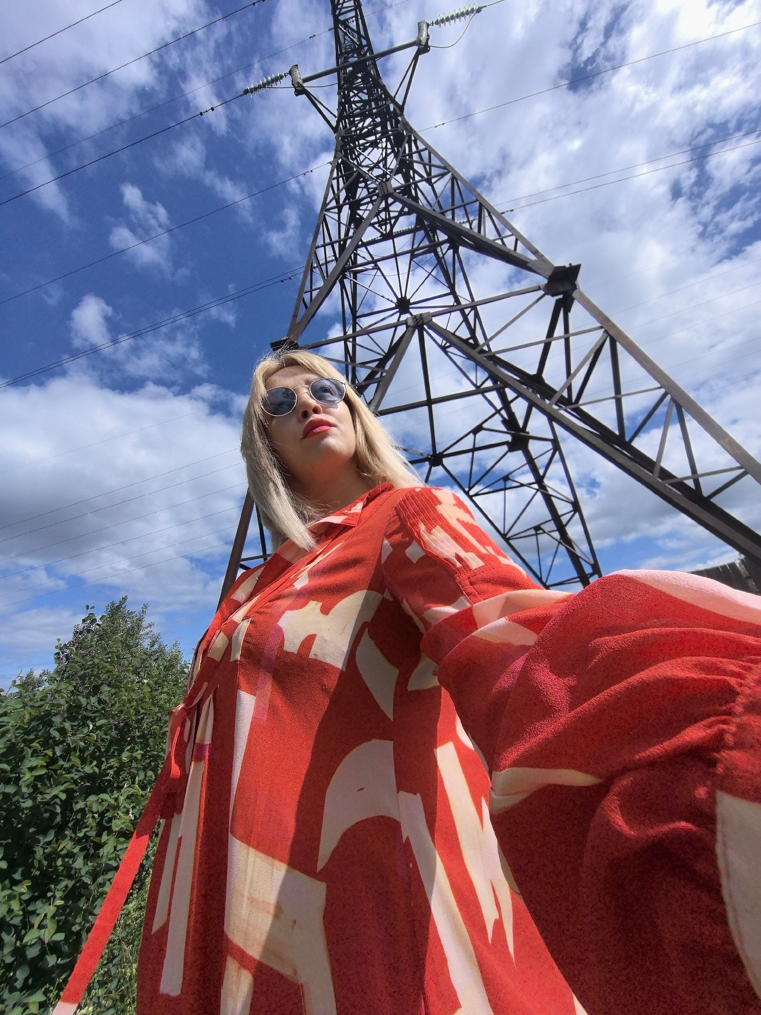 A woman with blonde hair and sunglasses stands in front of a tall power line tower under a blue sky with clouds. She is wearing a red and white patterned dress. The image appears to be taken outdoors in a rural or suburban area, possibly in Noviy, Russia, given the power line infrastructure and vegetation visible. The perspective is from a low angle, looking up towards the tower and the woman.
