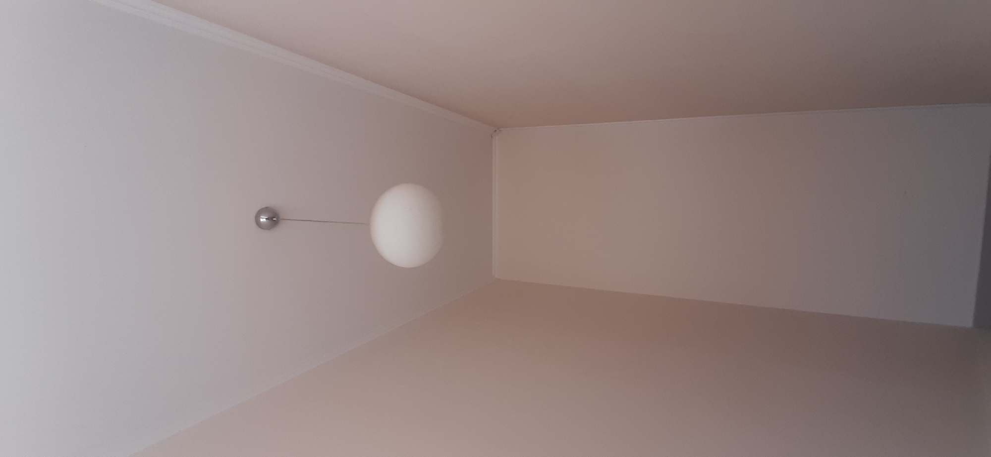 The media file captures a close-up interior shot of a room with light-colored walls and ceiling. A minimalist modern pendant light hangs from the ceiling. The light fixture consists of a small, metallic sphere attached to the wall by a thin wire, from which a larger, opaque white spherical lamp shade is suspended. The lighting suggests an indoor setting, likely during the daytime, as there are no strong artificial light sources visible and the ambient light is soft. The clean lines of the room and the simple design of the light fixture suggest a contemporary aesthetic. The scene appears to be a static view, with no activity taking place. The location context indicates Yessentuki, Russia, but there are no specific visual cues in the image to confirm this. The overall mood is serene and uncluttered. No text is visible in the image.