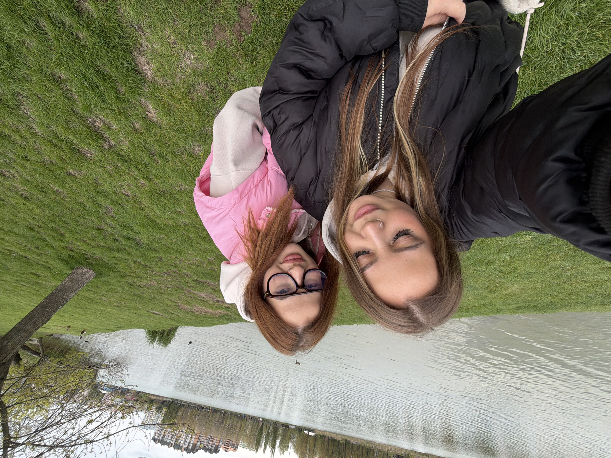 This overhead selfie captures two young women relaxing outdoors by a body of water, presumably in Krasnodar, Russia.
The woman positioned on the left (when viewed upright) has reddish-brown hair and is wearing glasses. She is dressed in a light pink hooded jacket over a lighter-colored top, looking up at the camera with a neutral or subtly smiling expression. The woman on the right has long brown hair with blonde highlights and sports a nose piercing. She is wearing a black puffer jacket over a light top, and her eyes are closed or partially closed, revealing a gentle smile.
Both subjects appear to be lying on a grassy bank. To their side, a calm body of water, likely a lake or river, stretches into the distance, showing faint ripples on its surface. Along the far bank of the water, several buildings and a line of trees are visible, indicating an urban or suburban environment. A tree trunk is also present on the near grassy bank. Small birds, possibly ducks, can be spotted on the grass near the water's edge and in the water itself.
The lighting suggests an overcast daytime, providing a soft and diffused illumination. The overall scene conveys a sense of tranquility and relaxation. No visible text is present in the image.