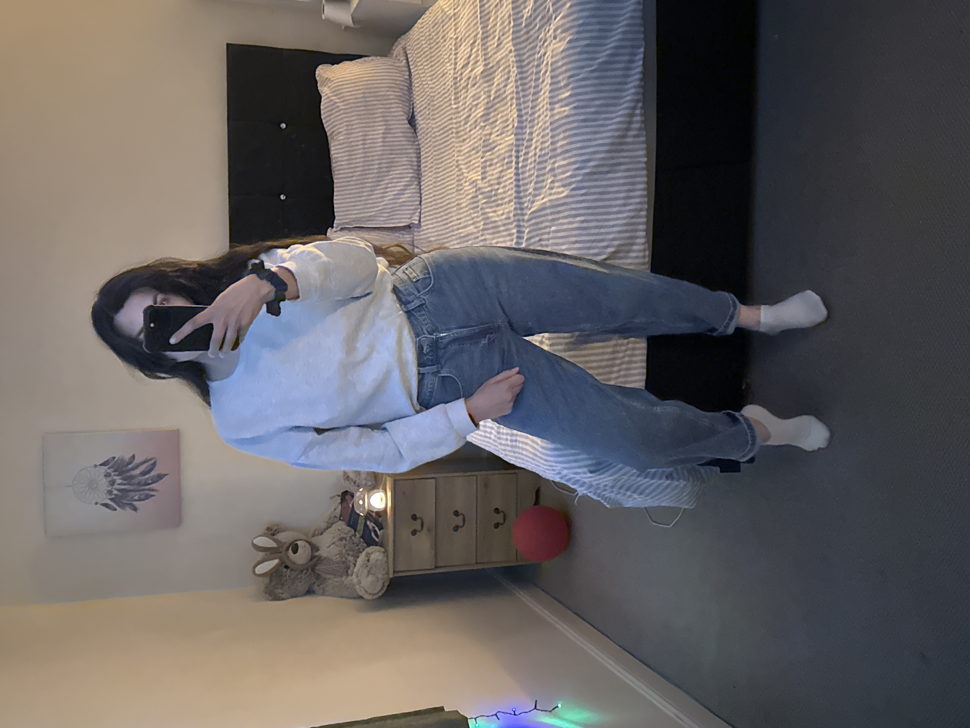 The image depicts a young woman in a bedroom setting. She is holding a smartphone and taking a photo of herself. She is wearing a light-colored sweatshirt and jeans, and white socks. The background includes a bed with striped bedding and a black headboard, a bedside table with drawers, a teddy bear, a red ball, and wall decorations (a dreamcatcher-like artwork). Christmas lights are visible on the floor. There's no clear indication of this image being from the city of Great Yarmouth.