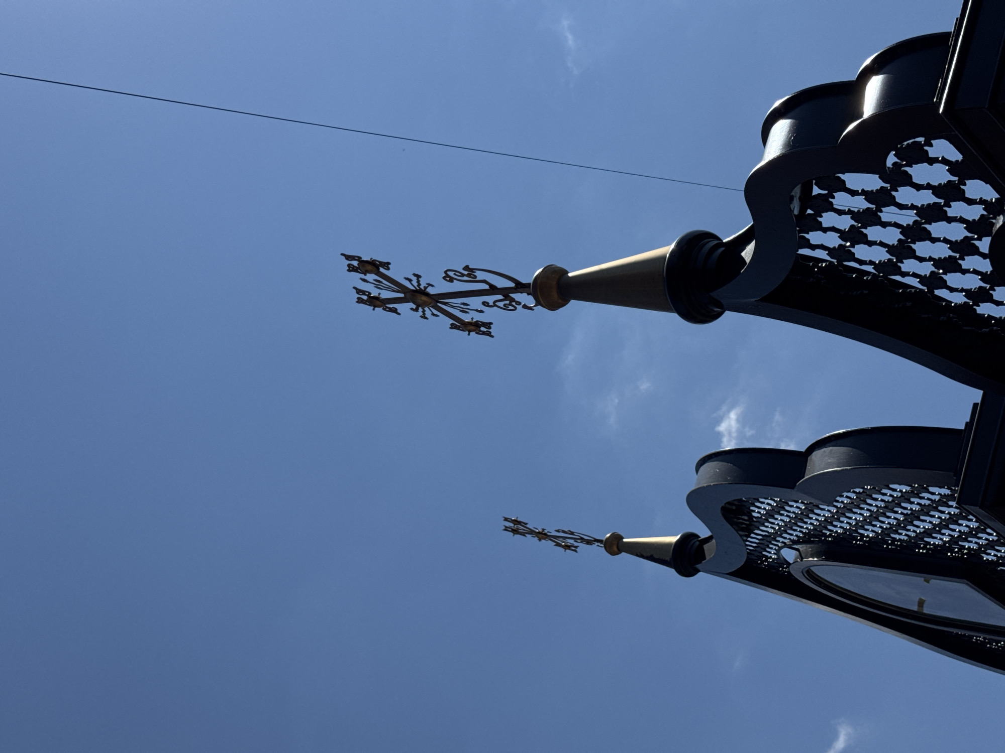 An upward angle shot of two ornate crosses atop dark architectural elements, set against a clear blue sky. The crosses are detailed and appear to be made of metal, possibly gold-colored. The architectural elements below them are dark, possibly black or very dark metal, with decorative cut-out patterns. A thin power line stretches diagonally across the upper right portion of the sky. One of the architectural elements features a circular clock face. The scene suggests a building with a prominent roofline or spire. The location is Unknown, Unknown.