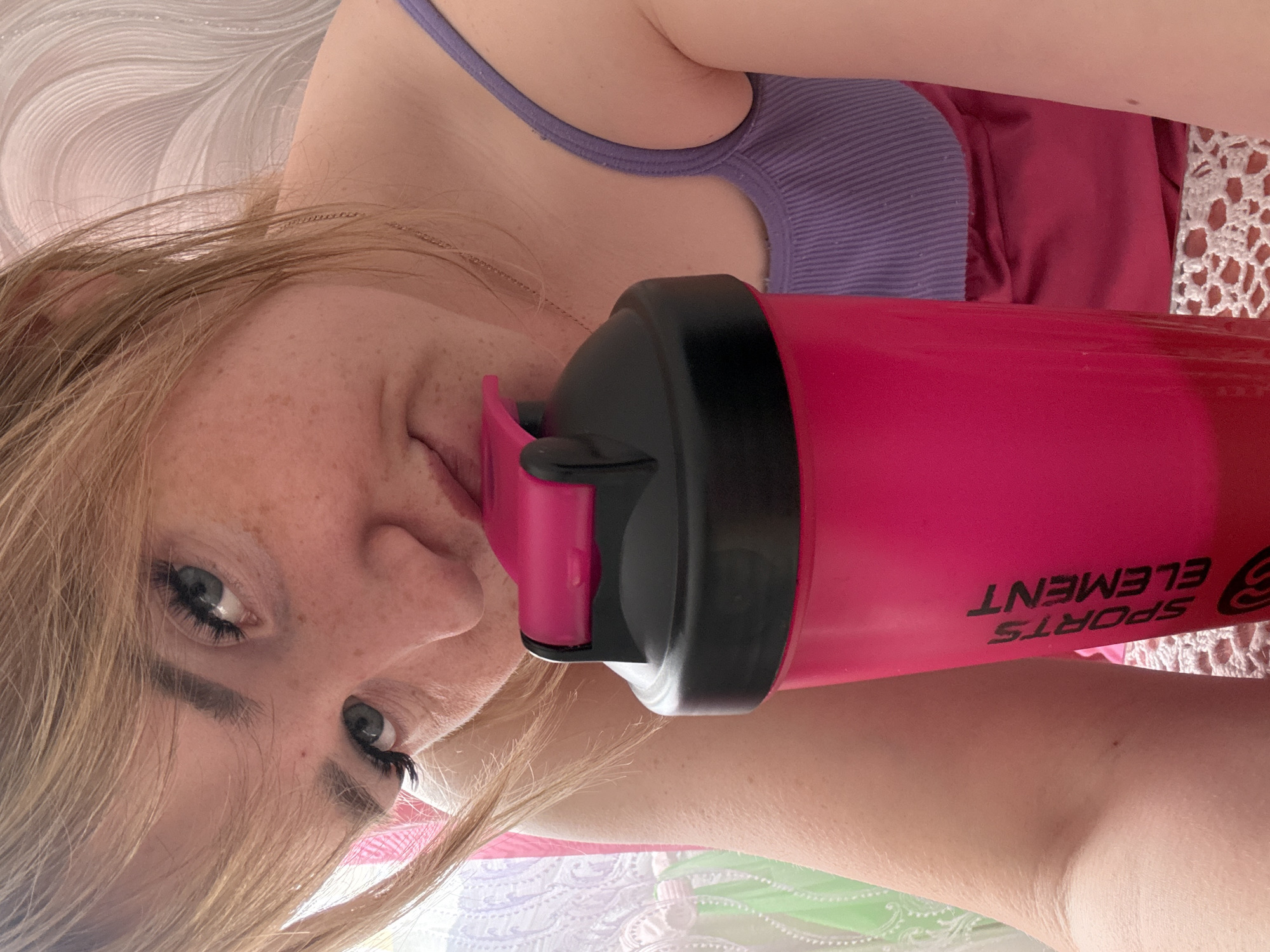 A close-up, rotated image features a fair-skinned person with freckles and light brown hair, holding a pink and black "Sports Element" branded water bottle. The person is wearing a lavender tank top under a pink shirt, and a thin necklace is visible. The background includes a light-colored patterned surface, possibly fabric. The location and city are not discernible from the image.