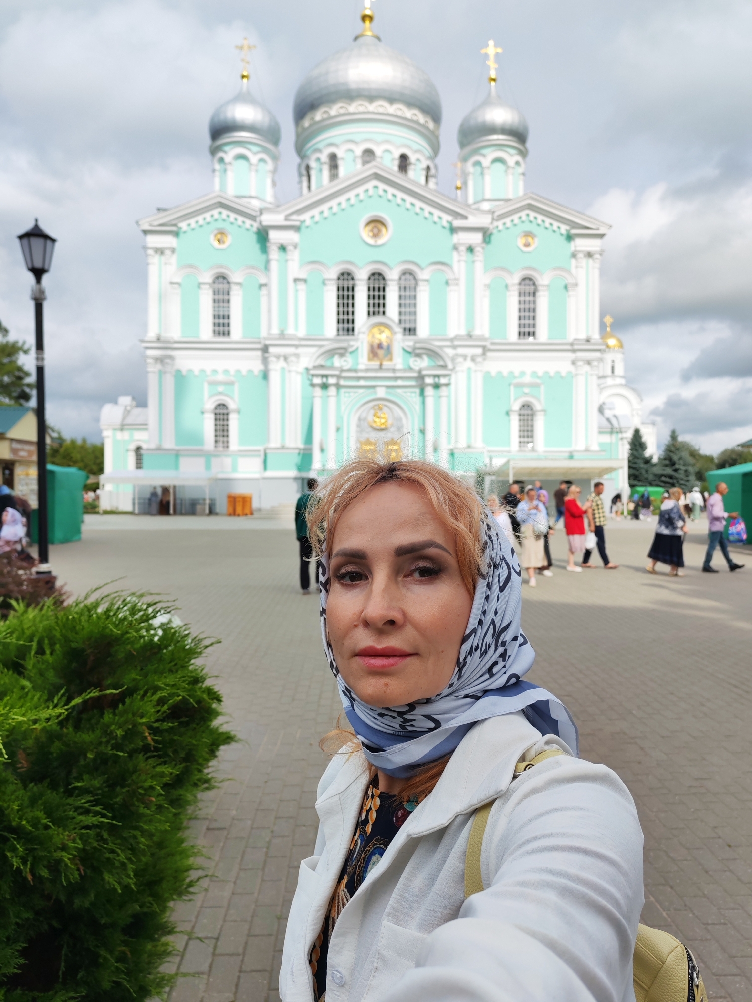 A medium shot captures a woman taking a selfie in front of the Trinity Cathedral in Diveyevo, Russia. The woman, with blonde hair and wearing a white jacket, a colorful scarf, and a backpack, is positioned in the foreground, with her face filling a significant portion of the left side of the frame. Behind her, the teal and white facade of the majestic Trinity Cathedral dominates the background, its silver domes glinting under a cloudy sky. The ground is paved with interlocking brick-like stones. Several people are visible in the mid-ground, walking in front of the cathedral, their faces indistinct. A lamppost stands to the left of the frame, near a lush green shrub in the foreground. The overall scene depicts a moment of personal reflection and admiration of the architectural landmark.