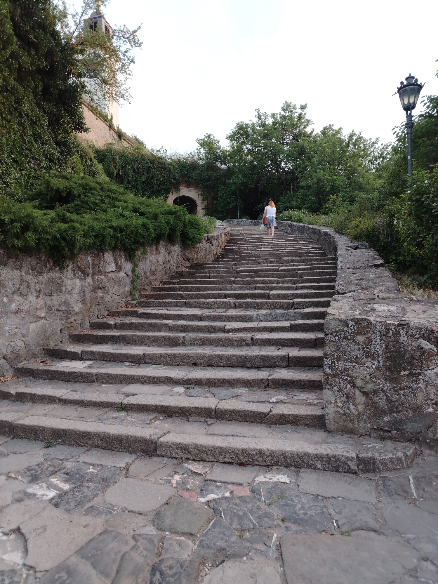 An outdoor, upward-angle shot shows a person walking up a wide stone staircase. The staircase is made of large, rough-hewn stones, with a stone wall bordering the right side and a retaining wall covered in green foliage on the left. The person, who appears to be a woman with red hair, is wearing a white top and a striped skirt and is carrying a bag. She is walking away from the camera, towards a tunnel-like opening visible in the distance at the top of the stairs, framed by trees. The scene is set within the Petrovaradin Fortress in Petrovaradin, Serbia, with glimpses of a clock tower peeking out from behind the foliage on the left. A vintage-style lamppost is visible on the right side of the image, amidst the greenery. The sky above is overcast. The foreground shows a paved pathway made of irregular stones.
