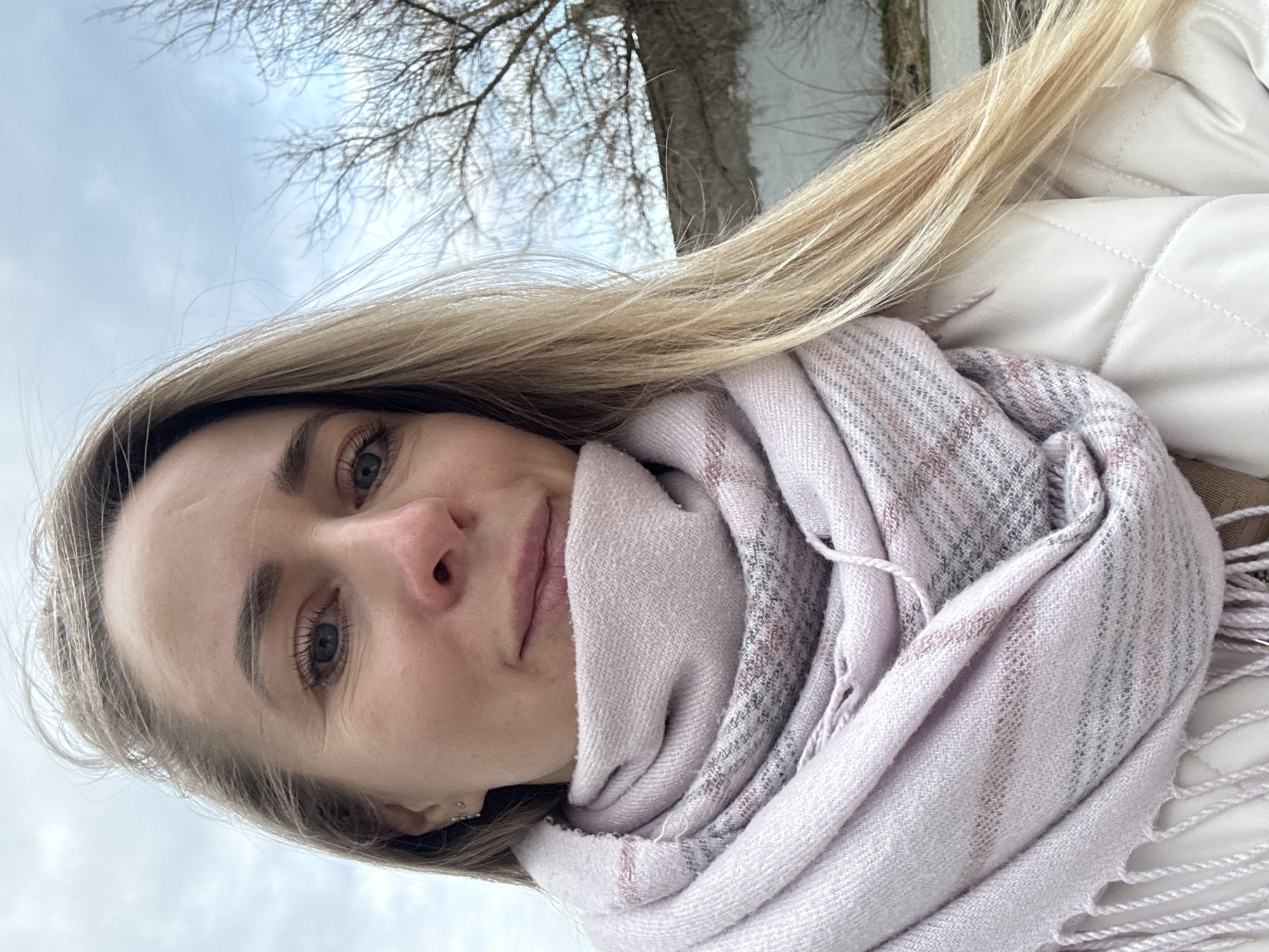 A woman with long, blonde hair and light skin is taking a selfie. She is wearing a light-colored quilted jacket and a light pink scarf wrapped around her neck. Her eyes are blue. The background shows a pale, overcast sky, bare tree branches, and a gray wall, potentially a building or embankment. The location is possibly the city of Chaikovsky, Russia.