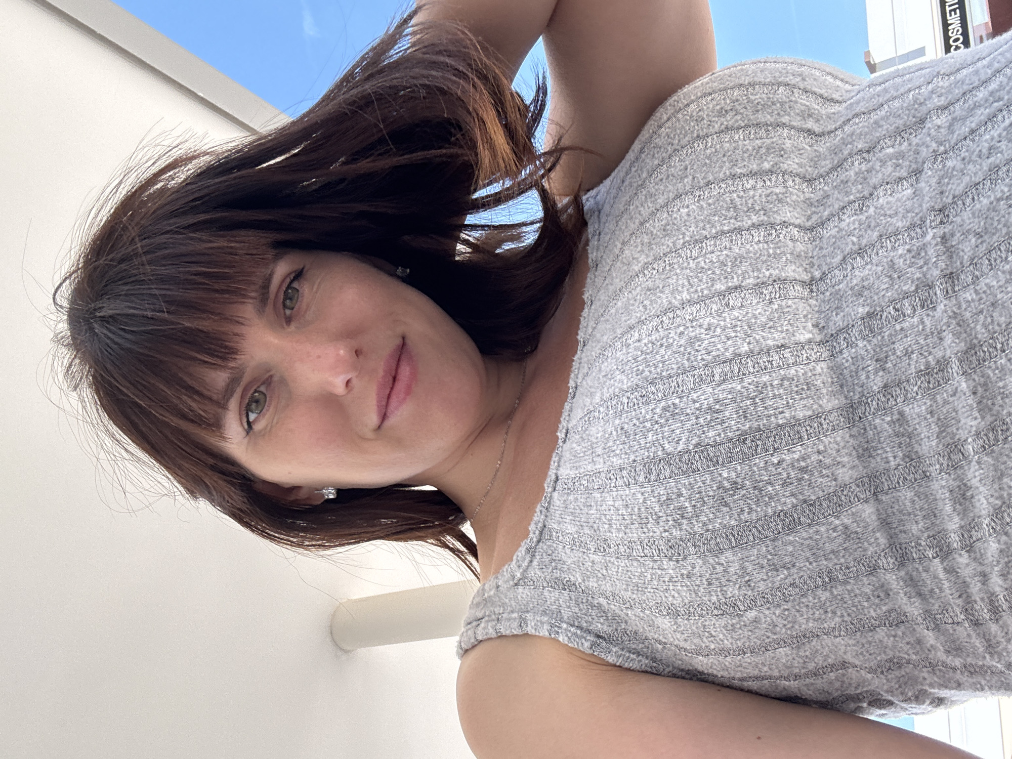 A woman with brunette hair and bangs is looking down at the camera. She is wearing a gray, sleeveless top. The sky is blue in the background. "COSMETIC" is partially visible on a building sign behind her. The exact location within the city of Pomezia, Italy, cannot be determined from the image.