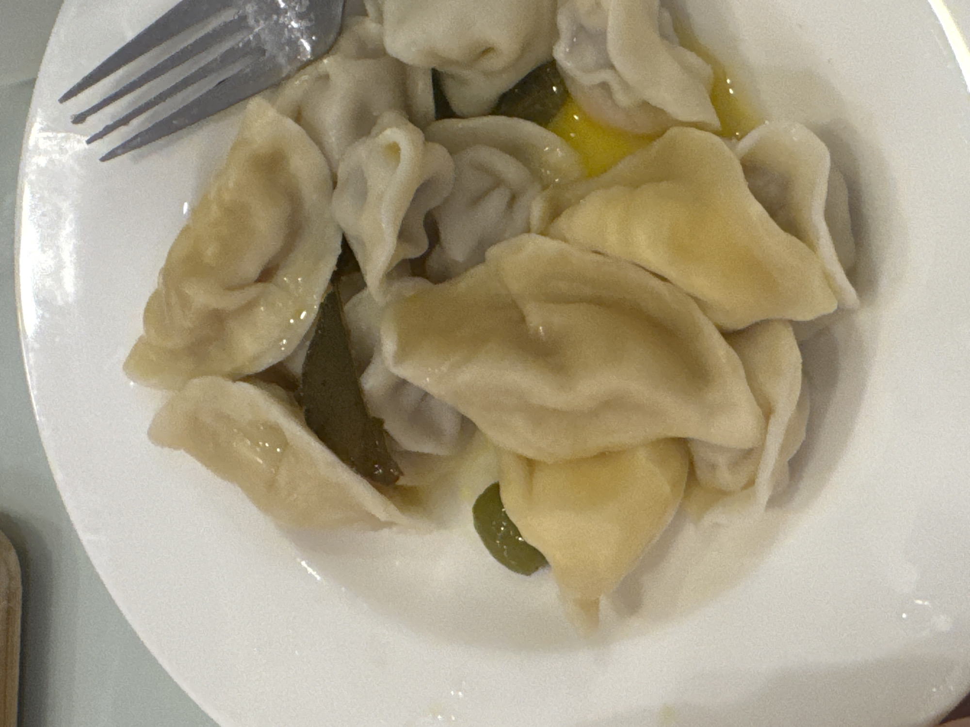 A top-down view shows a white plate containing a pile of dumplings, some of which are glistening with what appears to be melted butter or oil. A silver fork rests on the upper left edge of the plate. Several dark green, oblong objects, possibly pickles or olives, are interspersed among the dumplings. The location of the scene is unknown.