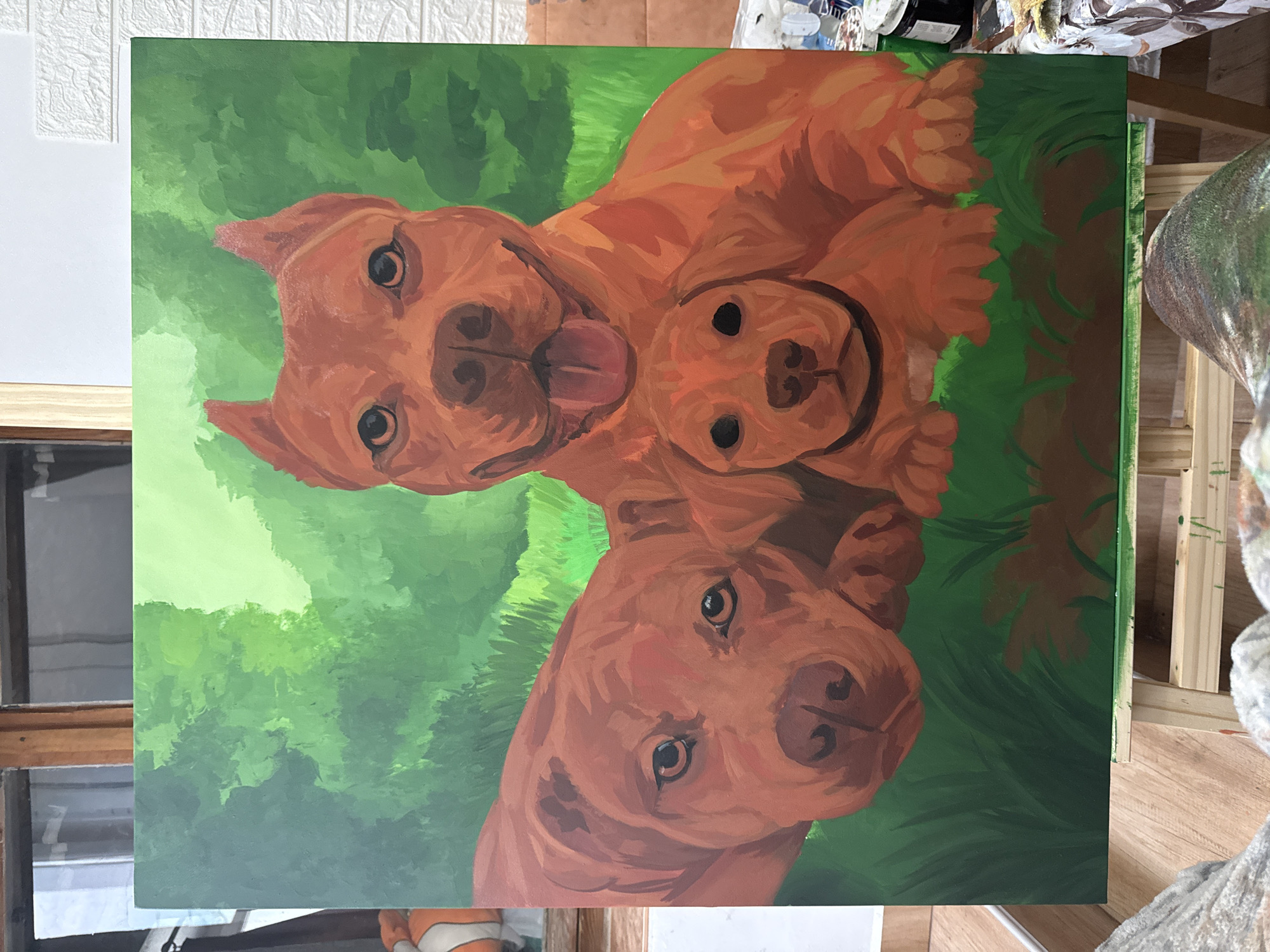 The image depicts an acrylic painting on canvas of two adult pit bulls and one puppy. The dogs are rendered in shades of brown, with the adult dogs having detailed facial features and the puppy simplified. The background is a blurry mix of green, suggestive of a grassy or wooded environment. The painting is set on a wooden easel. A window and white wall are visible in the background. The image was likely taken in the Generalovo, Bulgaria.