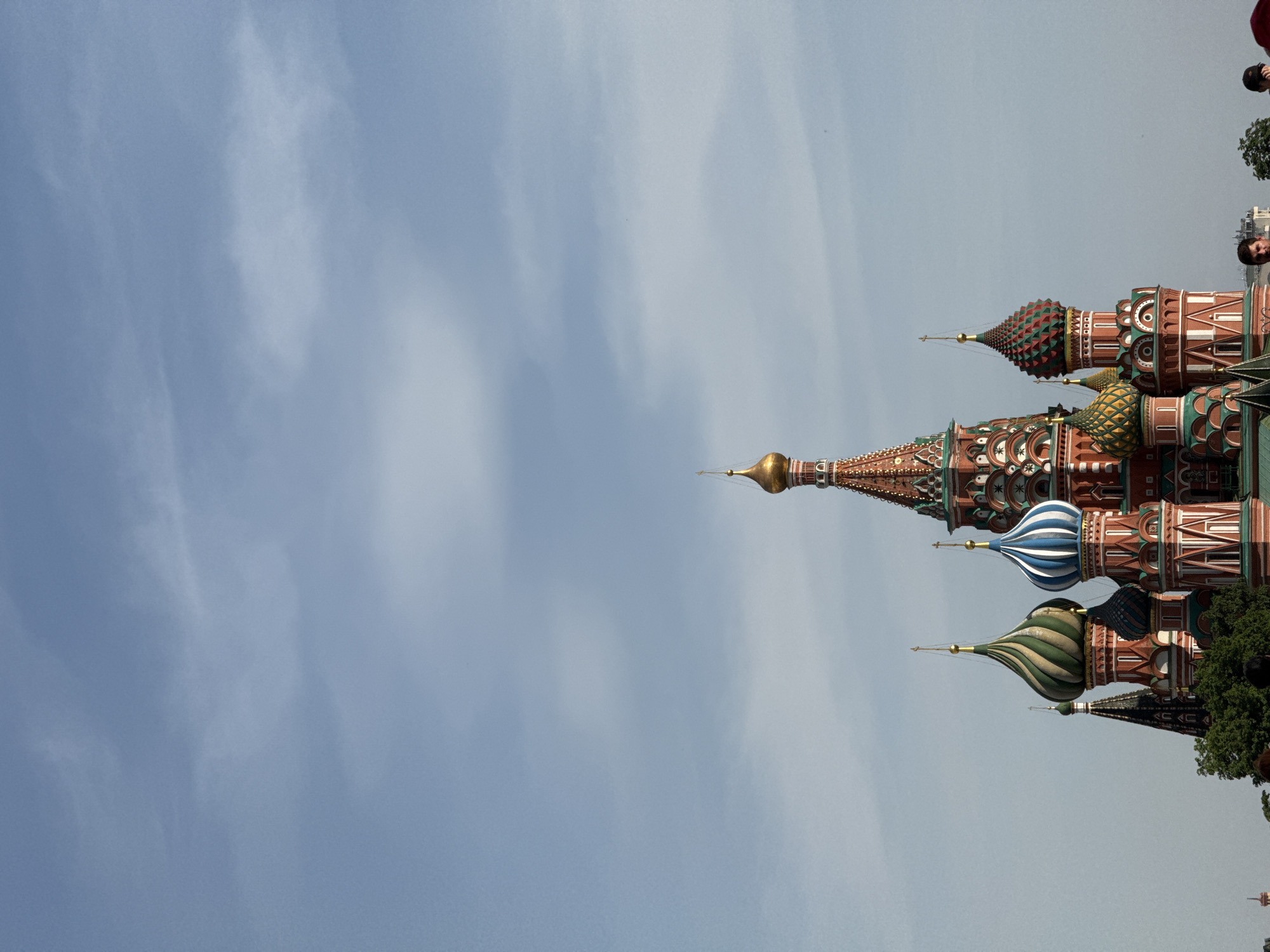 The image shows a partial view of St. Basil's Cathedral in Moscow, Russia, with its distinctive colorful onion domes against a light blue sky with subtle cloud formations. The domes display varied patterns and colors, including green, blue, and red, atop elaborately decorated brickwork. Partial views of people are visible in the top right corner of the image.