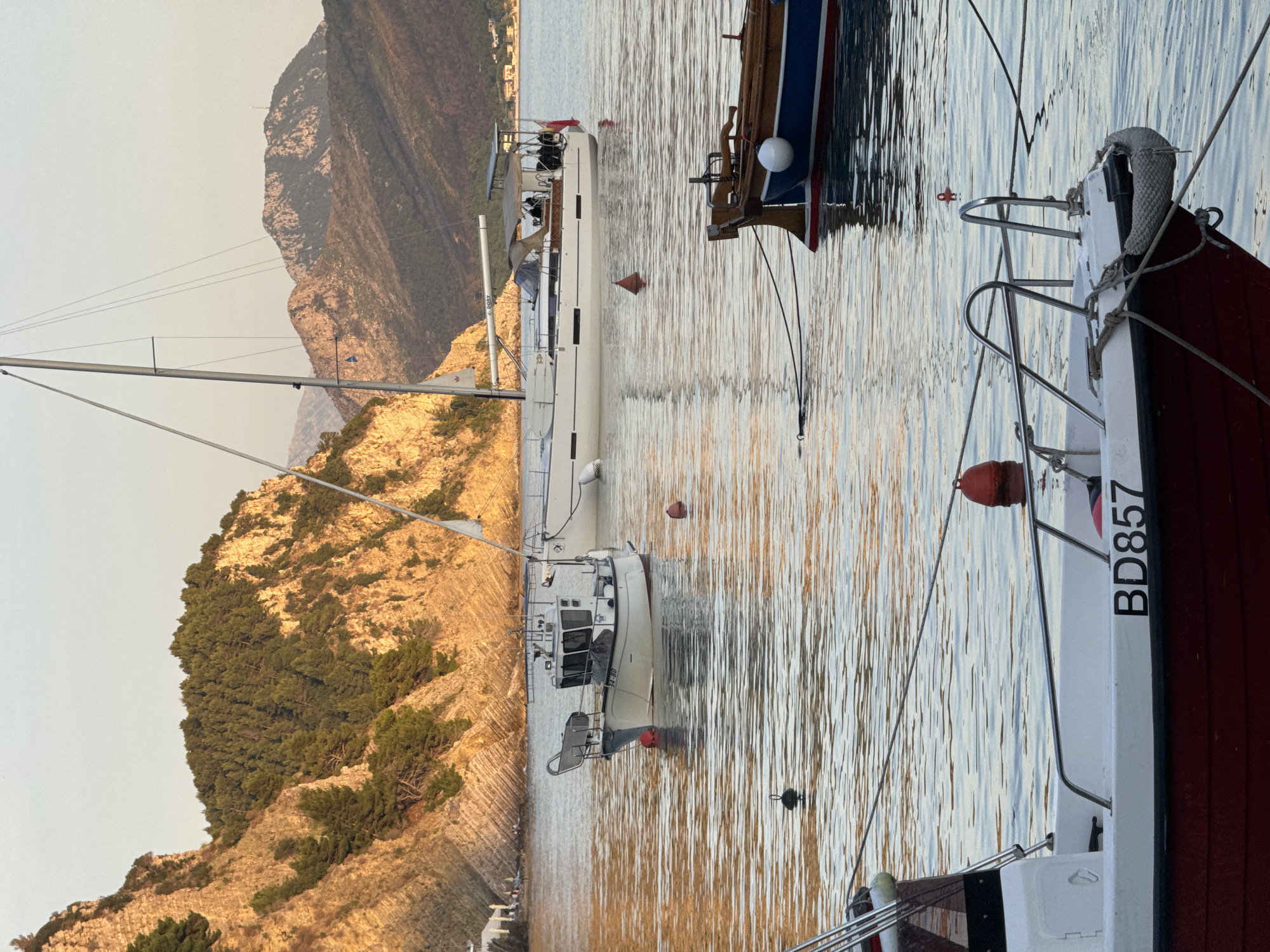 The image depicts a harbor scene in Petrovac centar, Montenegro, at what appears to be either sunrise or sunset given the warm lighting. The primary subjects are three boats of varying sizes moored in the calm water. A large, white sailboat occupies the center of the frame, flanked by two smaller boats on either side. The boat on the right has the letters "BD857" visible on its side. Behind the boats, the coastline rises dramatically, featuring a steep, rocky cliff covered with trees. The sky is a hazy color, consistent with the time of day. The water surface reflects the colors of the sky and the surrounding landscape.