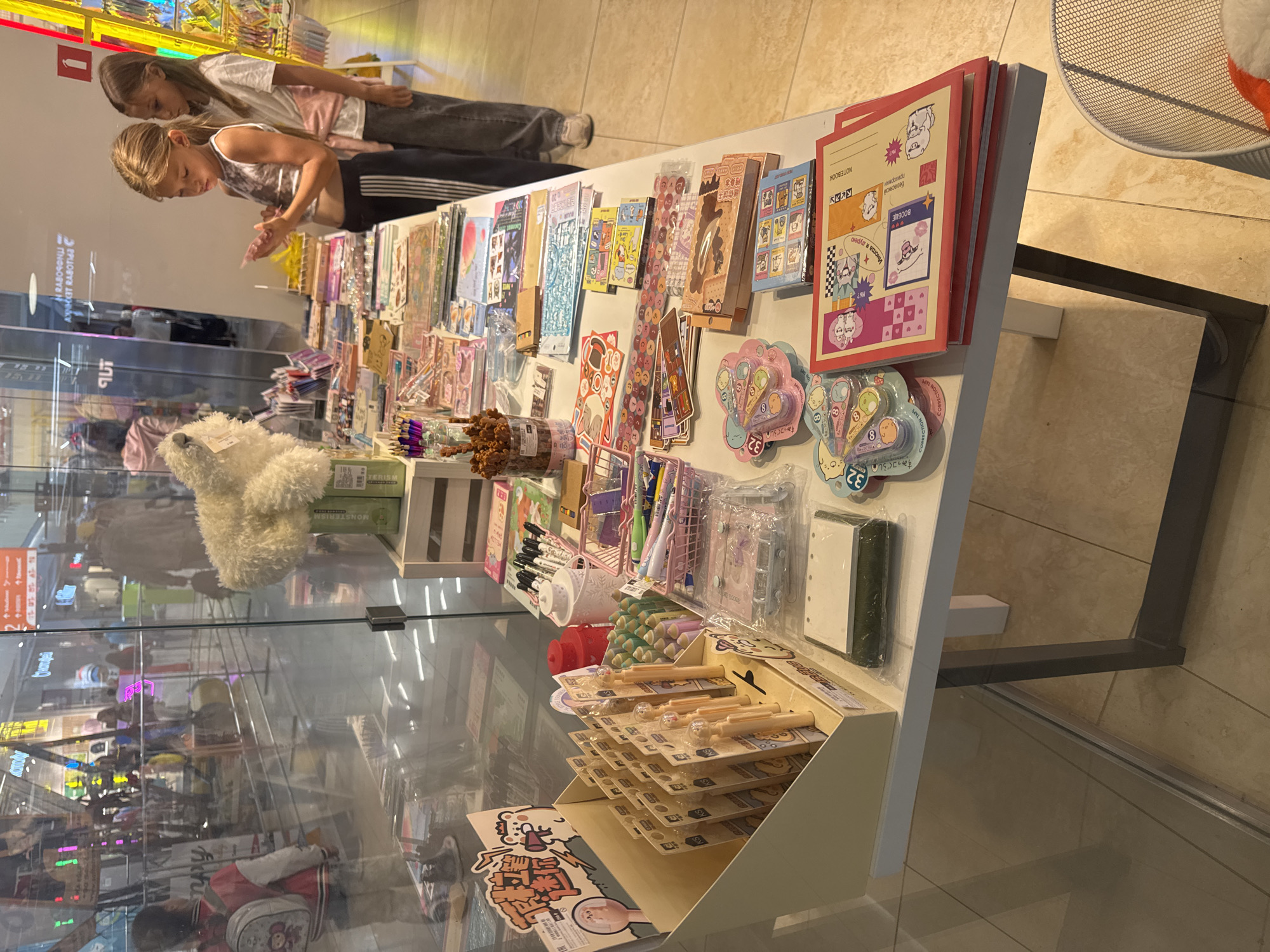 This is a medium-angle, eye-level, outdoor full shot of a retail display of stationery and stationery accessories in a brightly lit shopping mall. Two young girls, likely sisters, are browsing the display. The girl on the left is wearing a white tank top and black athletic pants and is examining a small item in her hands. The girl on the right is wearing a white t-shirt and gray pants, with a pink cardigan tied around her waist, and is looking at the display. The display table is covered with various items, including stickers, pens, notebooks, erasers, and other small stationery items, neatly organized in boxes and baskets. In the background, other shoppers and store displays are visible through a glass partition. The flooring is tiled, and the overall atmosphere suggests a commercial setting. The image's metadata indicates it was taken in Ufa, Russia.