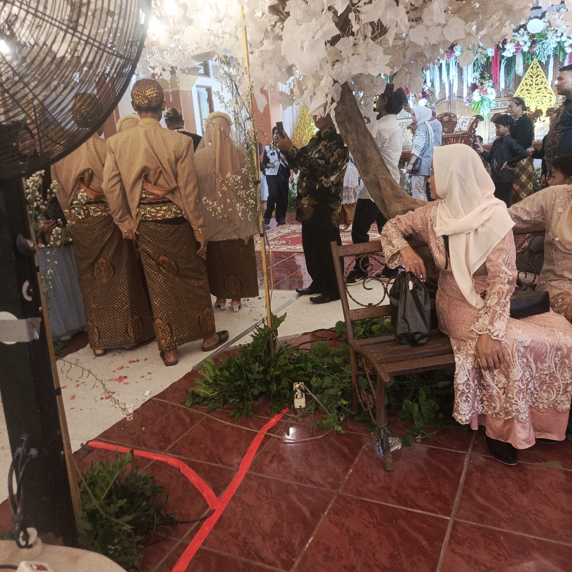 This is a medium shot, eye-level, indoor photograph taken at a wedding reception, likely in South Jakarta, Indonesia. The scene is decorated with white artificial flowers cascading from the ceiling and down a wooden tree-like structure on the right. A large fan is visible on the left, obscuring part of the background.
In the foreground, to the right, a woman wearing a light pink traditional dress and a white headscarf sits on a rustic wooden bench. She is holding a black bag on her lap and has her hands resting on her knees. To her right, another woman in a similar pink dress is partially visible.
In the midground, two men in traditional Javanese attire, including tan blazers and patterned brown sarongs, are standing. They appear to be part of the wedding party. Behind them, several other guests are seen, some in traditional Islamic attire with headscarves. One man in a dark patterned shirt is taking a photo with his phone.
The floor is tiled with reddish-brown tiles, and red tape is laid out on the floor in a cross pattern. Green foliage is arranged around the base of the decorations and along the floor. In the background, there are more wedding decorations, including a tiered structure with floral arrangements and golden accents. The overall atmosphere suggests a festive, cultural event.