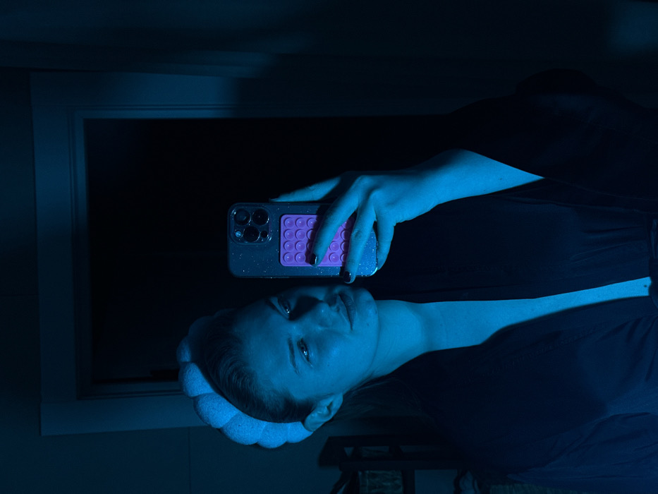 A woman with a white padded headband is taking a selfie in a dark room illuminated by a blue light. She is holding a phone with a clear, glittery case and a purple "pop it" fidget accessory. The light casts shadows, highlighting her face and neck, and illuminates a window frame behind her. Her black robe is visible.
