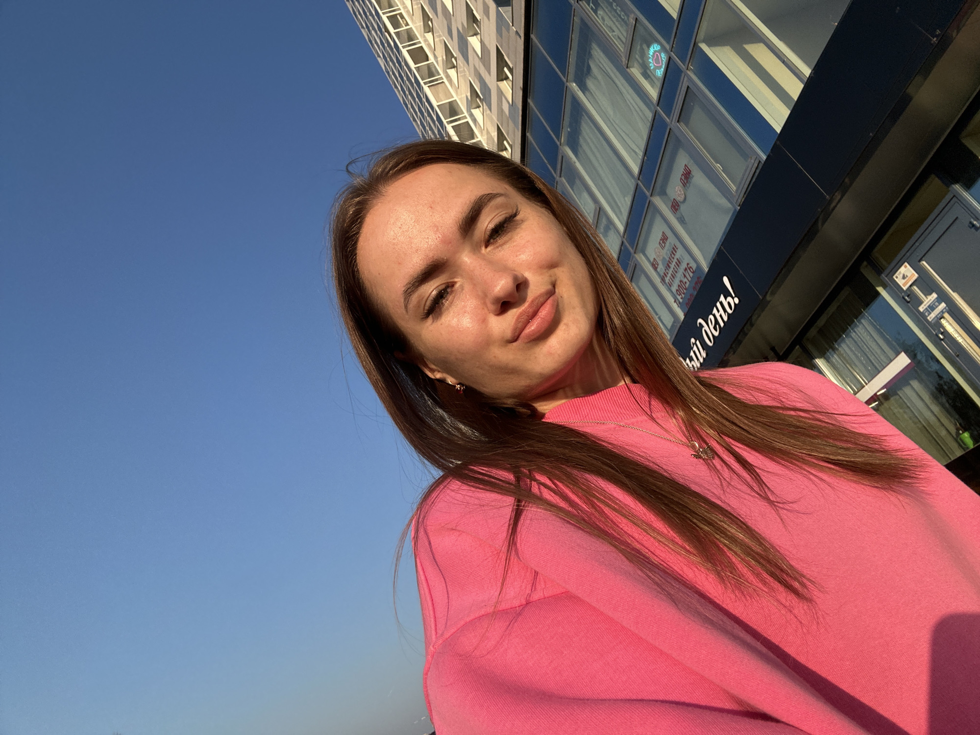 A young woman is captured in a selfie outdoors, with a bright blue sky dominating the upper left quadrant of the image. She is wearing a vibrant pink sweatshirt and a delicate necklace with a butterfly pendant. Her long, brown hair is styled down, framing her face. In the background, a modern building with large windows and a visible sign featuring Cyrillic text stands tall. The lighting suggests it is daytime, possibly late afternoon, as evidenced by the warm glow on the woman's skin and the soft shadows. The scene appears to be in an urban environment, indicated by the presence of the building.