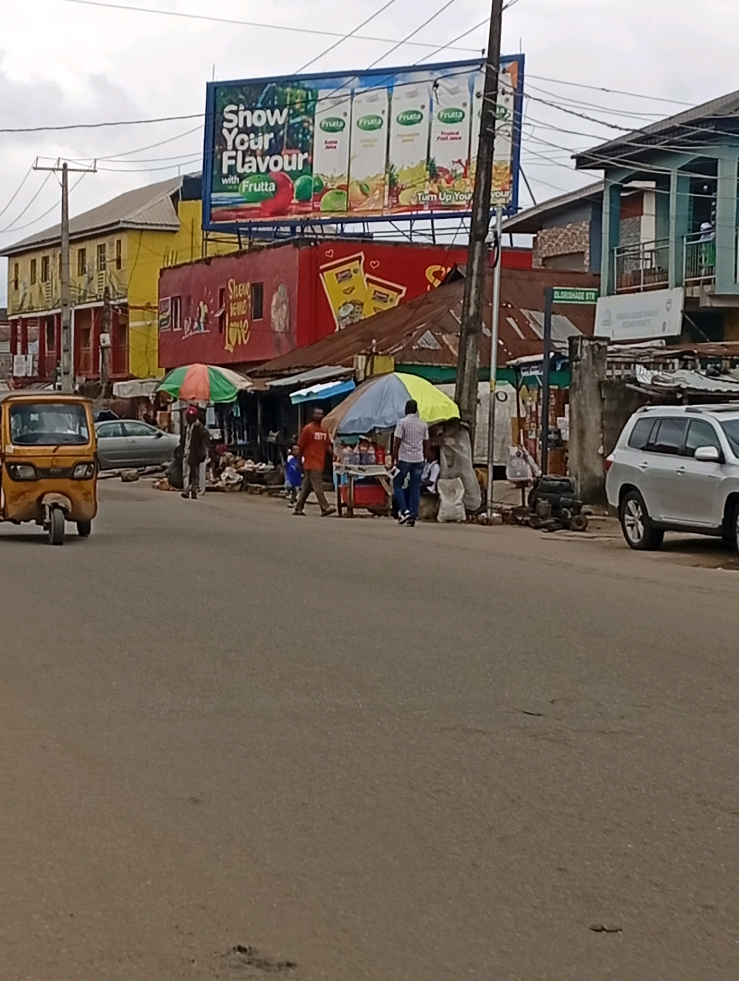 The image depicts a street scene in Akowonjo, Nigeria, with a focus on commercial activity. In the foreground, a paved road shows some wear and tear. To the left, a yellow three-wheeled auto-rickshaw is parked. Further down the street, several stalls are set up under umbrellas, with people engaged in commerce. There is a silver SUV parked on the right side of the road.
Dominating the background is a large billboard with the slogan "Show Your Flavour" and images of juice cartons and fruit. Beneath the billboard, a red building with "Show Some Love" written on it is visible. The architecture suggests a bustling urban environment with a mix of older and newer structures. Power lines are strung across the sky. The overall atmosphere appears to be that of a busy market or commercial area.