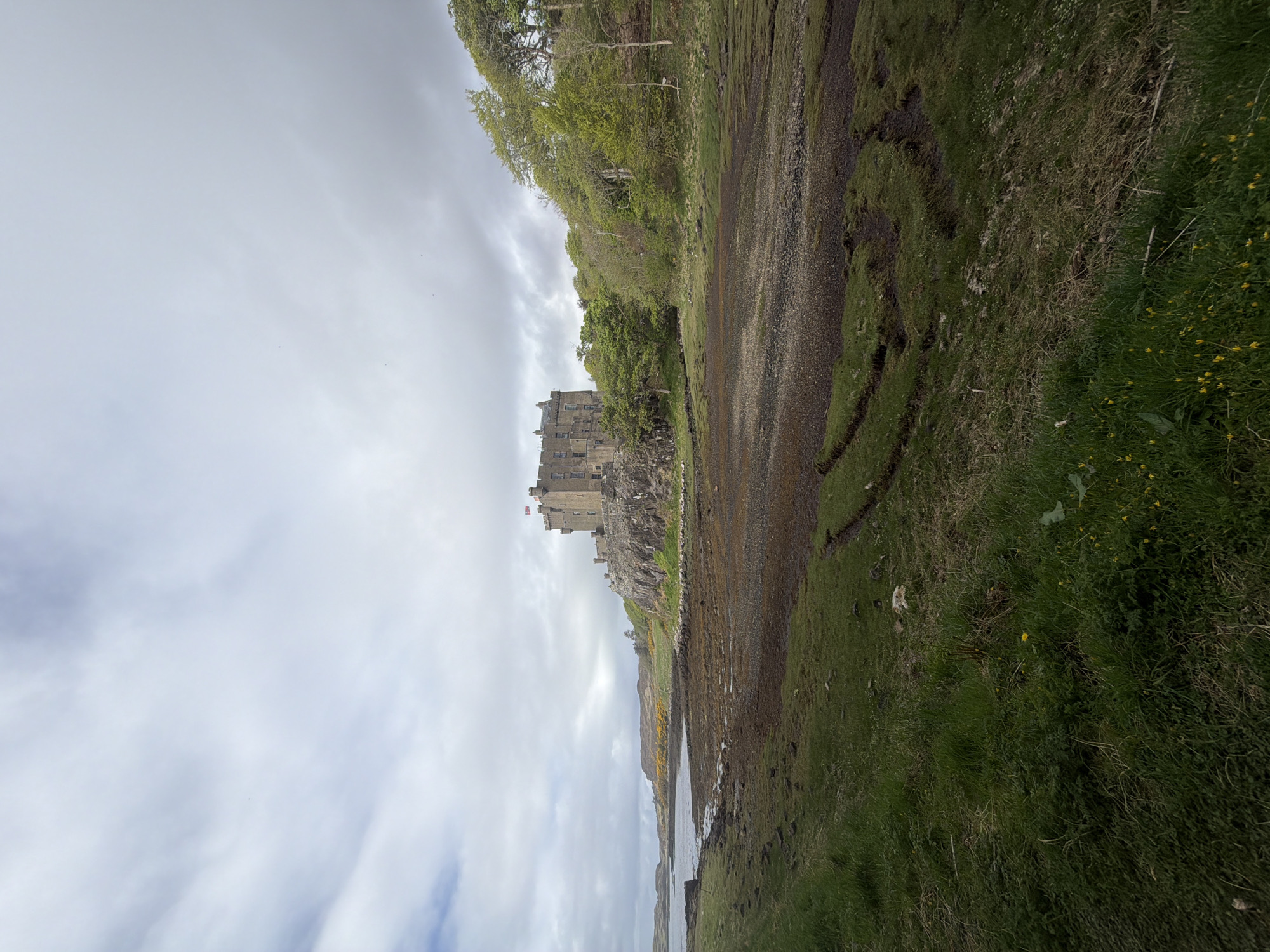 The image shows Eilean Donan Castle, a 13th-century castle situated on a small tidal island in Loch Duich, in the Scottish Highlands. The castle is visible in the center of the frame, a formidable stone structure with turrets and a flag flying atop. The surrounding landscape is characterized by a dramatic coastline, with rocky shores, green hills, and calm water of the loch. The sky is overcast, with white and grey clouds dominating the scene. The perspective of the image suggests a ground-level view, possibly taken from a nearby shoreline.
