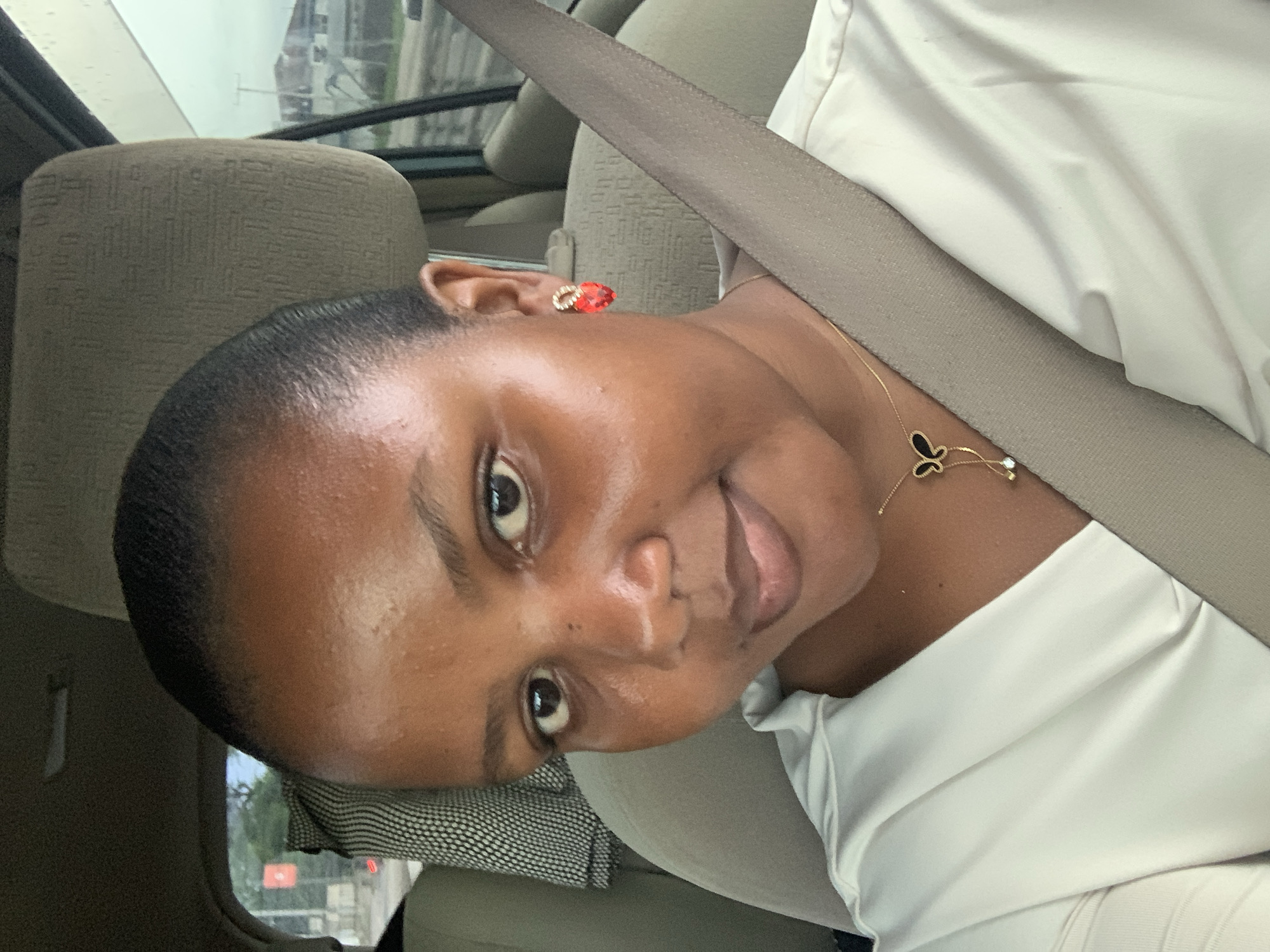 A close-up, rotated view depicts a dark-skinned woman wearing a white collared shirt and a seatbelt. She has short hair and is wearing red earrings and a gold necklace with a black butterfly pendant. The woman is inside a vehicle, evidenced by the upholstered seats. Through the window, buildings are visible, suggesting an urban setting. Given the prompt, the location is presumed to be Dar es-Salaam, Tanzania.