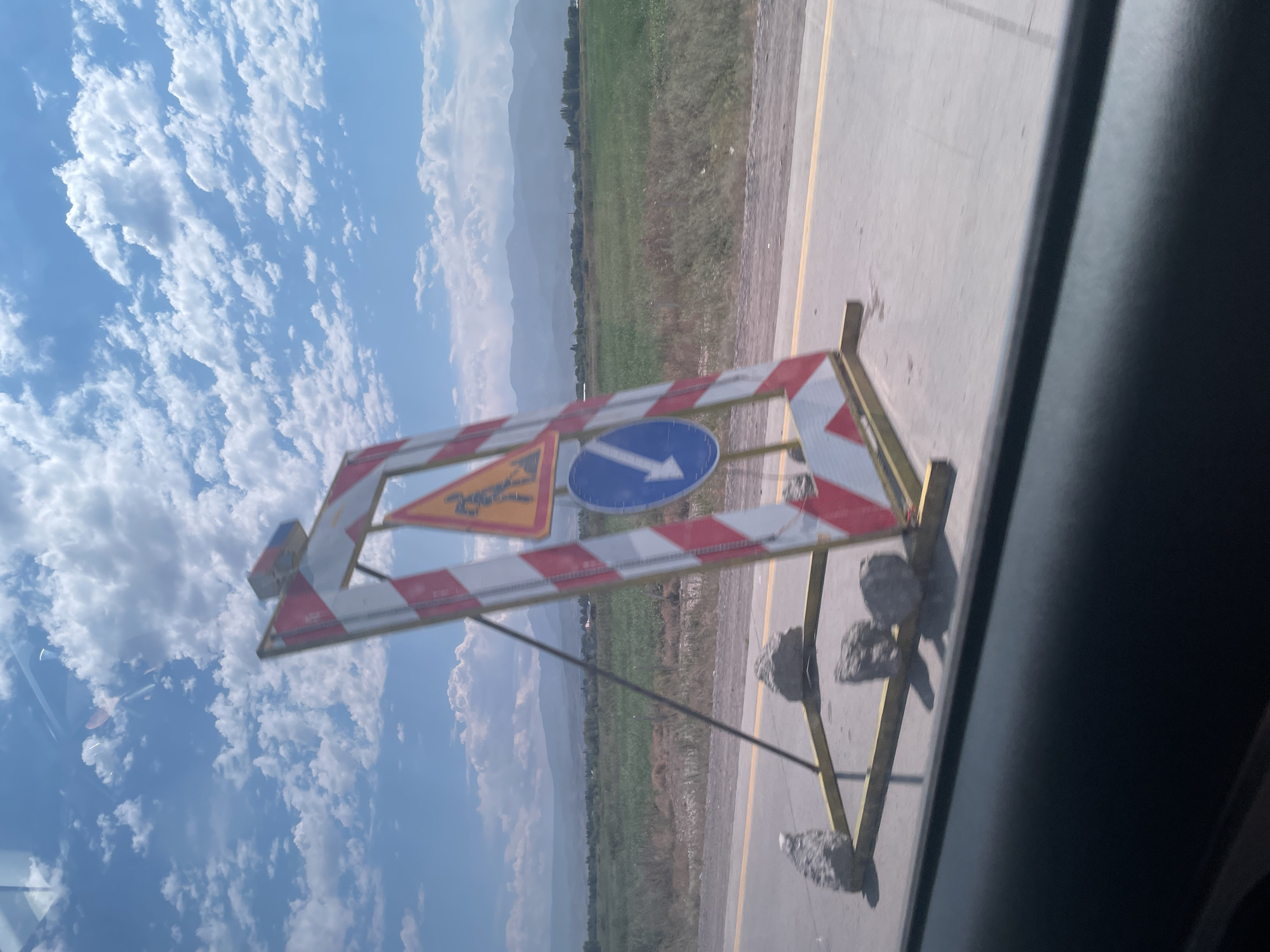 A construction barrier with red and white stripes stands on a paved road next to a yellow line, in the city of Qayrat, Kazakhstan. The barrier is supported by a metal frame and weighted down with rocks. A yellow triangular sign depicting a person with a shovel is attached to the barrier, as is a blue circular sign with a white arrow pointing down. The road is bordered by a grassy field with some dry vegetation and a line of trees in the distance. Mountains are visible on the horizon under a blue sky with scattered white clouds. The photo appears to have been taken from inside a vehicle, as indicated by the reflection in the upper left corner and the dark foreground.