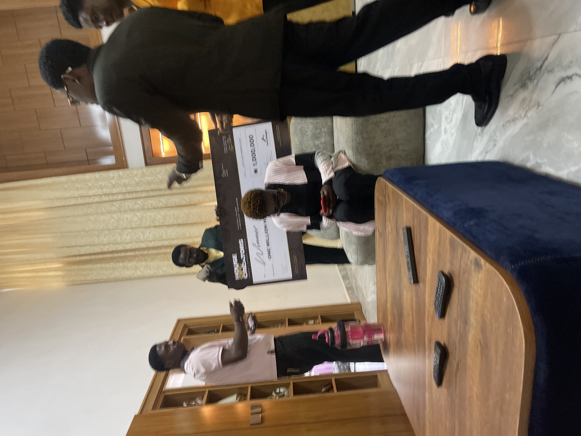 In a residential setting, likely within Gura Top, Nigeria, a group of individuals is gathered around a large novelty check. The check, predominantly dark with white text and graphics, prominently displays "HOUSE OF CREATORS" and a prize amount of "1,000,000". Two men are holding the check, one in a dark green suit and the other partially visible in a yellow top. Another man, wearing a pink shirt and black trousers, stands to the left, observing. Seated on a couch is a woman, dressed in a black and pink garment, looking down at her phone. In the foreground, a wooden coffee table with a dark blue base holds three remote controls. The background features cream-colored curtains and wooden paneling, suggesting an interior residential space. The lighting is warm, casting soft shadows.