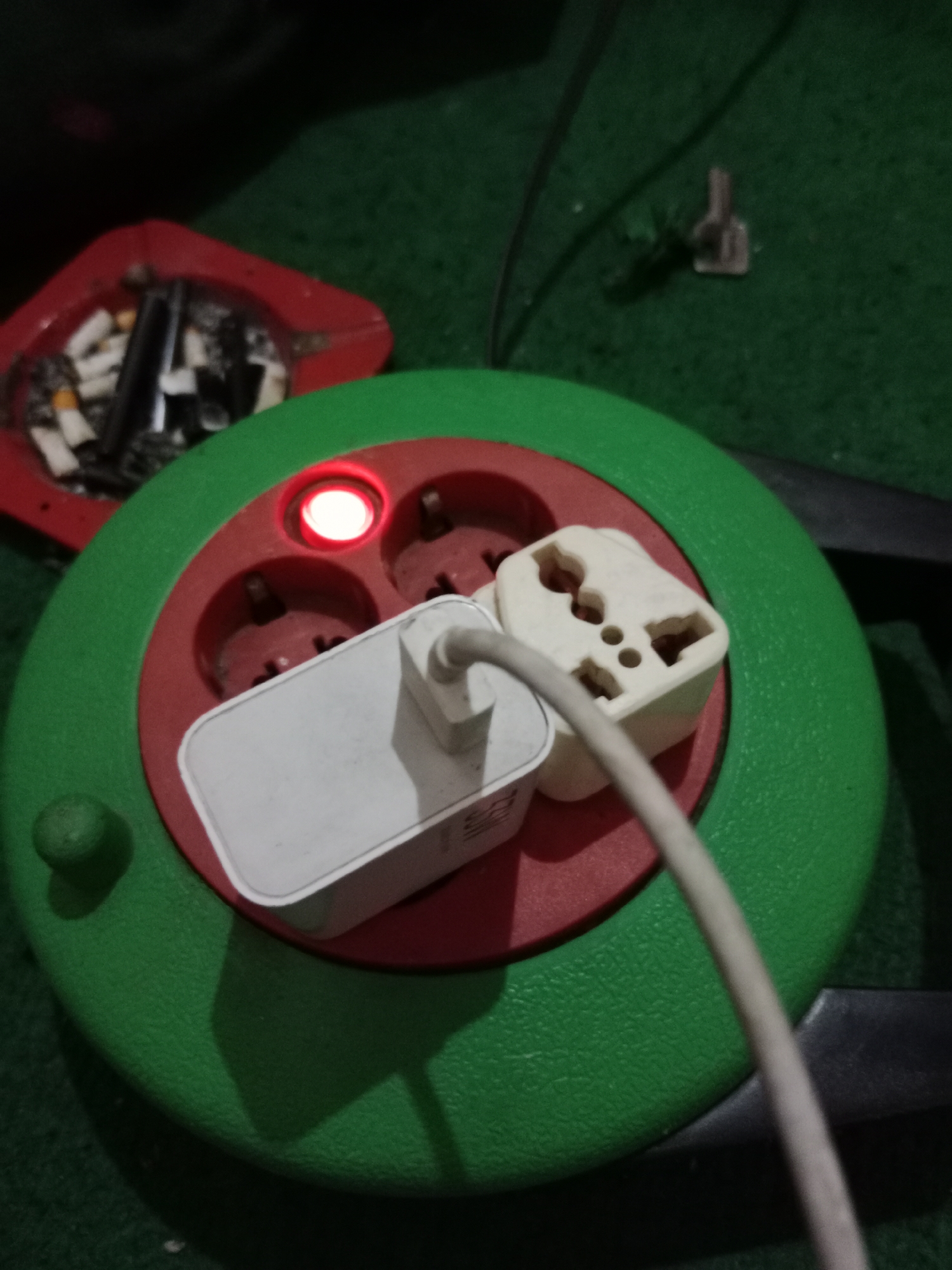In Ringinrejo, Indonesia, a close-up shot captures a green and red power strip on a dark green carpeted surface. The power strip features three outlets, one of which is illuminated by a small red indicator light. Two electrical adapters are plugged into the outlets. A white phone charger is connected to one adapter, and a grey cable extends from another adapter. To the left of the power strip, a red ashtray is visible, containing several cigarette butts and a black object. In the background, a small, light-colored object resembling a screw or nail rests on the carpet. The lighting is dim, suggesting an indoor environment.