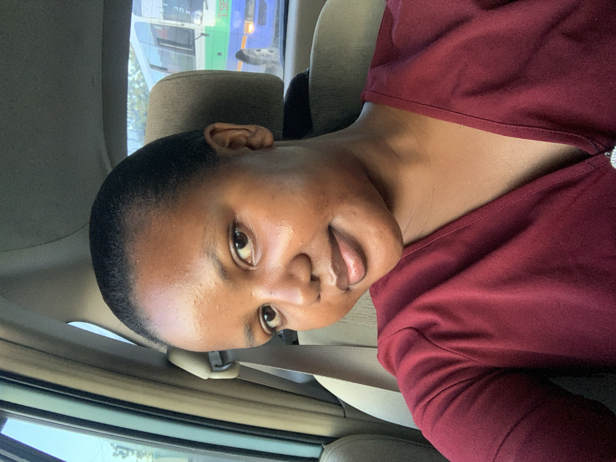 A person with dark skin and short black hair is inside a vehicle. The person is wearing a maroon-colored shirt and a seatbelt. Outside the car window, there is a bus with the words "TSEP" and "MNC" visible, suggesting the location is likely in Dar es-Salaam, Tanzania, based on the possible presence of the Dar es Salaam Bus Rapid Transit (BRT) system, which uses buses of this type. The interior of the vehicle appears to be light tan or beige.