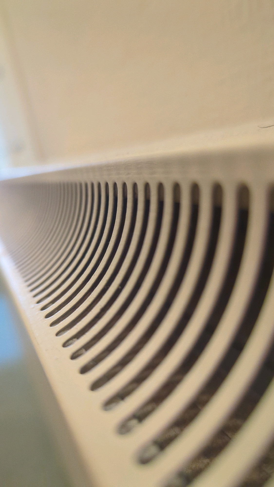 The image shows a close-up, low-angle shot of a beige-colored radiator grill. The grill consists of numerous curved slats, creating a series of openings that recede into the background. The lighting highlights the texture of the grill and casts shadows within the openings. The upper portion of the image is out of focus and appears to be a textured, light-colored surface, possibly a wall or ceiling. There are no people or discernible activities depicted. The location is stated to be Cornelscourt, Ireland, but this cannot be confirmed from the visual information alone.