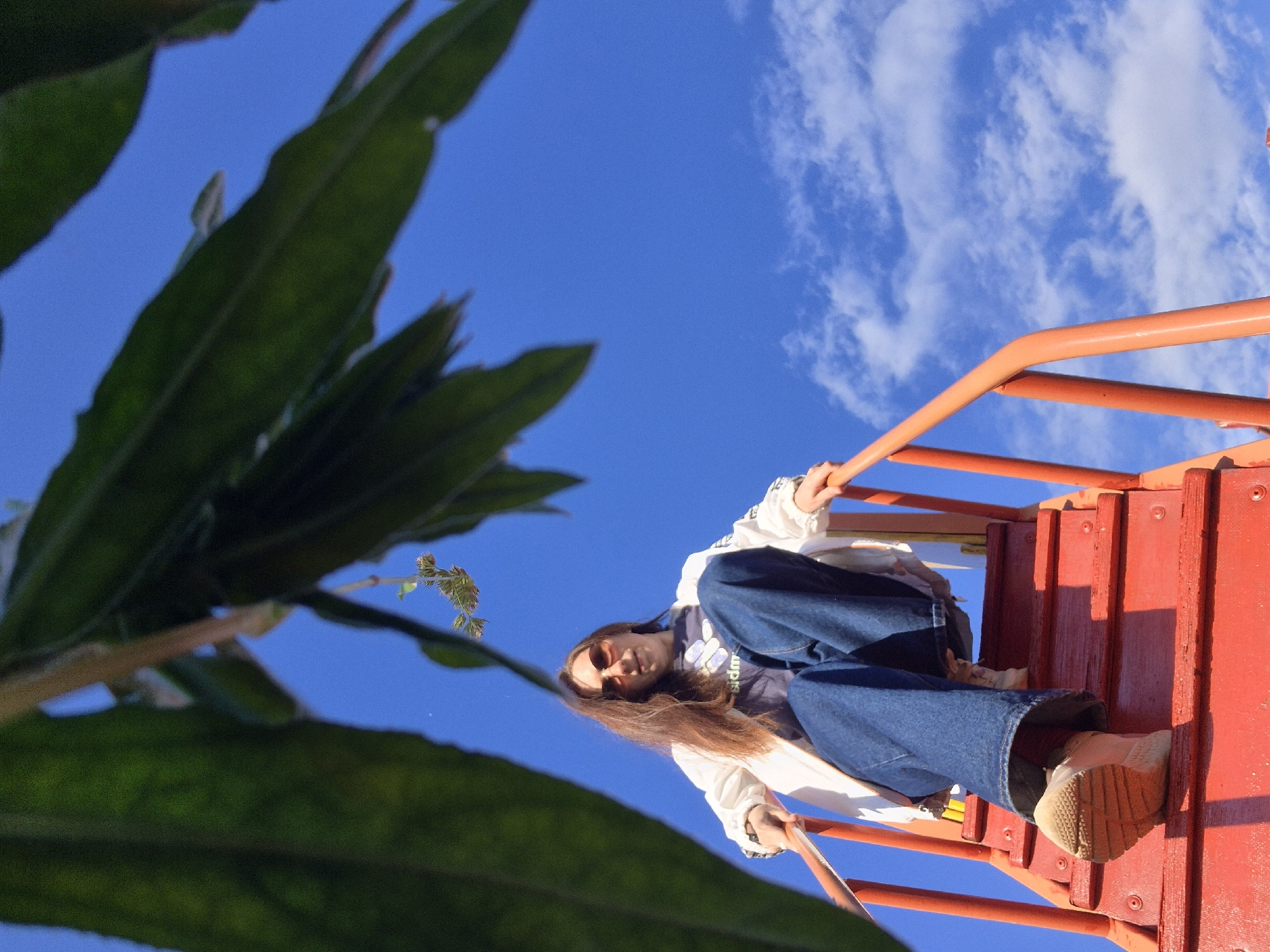 An upward-angled photograph captures a young person on a vibrant red playground structure against a clear blue sky dotted with scattered white clouds. The subject, who appears to be a young woman with long brown hair, wears sunglasses, a white jacket over a dark t-shirt, blue wide-leg jeans, and light-colored sneakers. She is seen sitting or leaning on the red steps of the playground, gripping bright orange railings, and looking towards the camera with a neutral expression.
Large, dark green leaves from a plant or tree occupy the left side of the frame, providing a natural foreground element and partially framing the scene. The bright, direct sunlight suggests a clear daytime setting. This scene is situated in Chaikovsky, Russia. A faint and indistinct white design or text is visible on the subject's dark t-shirt.