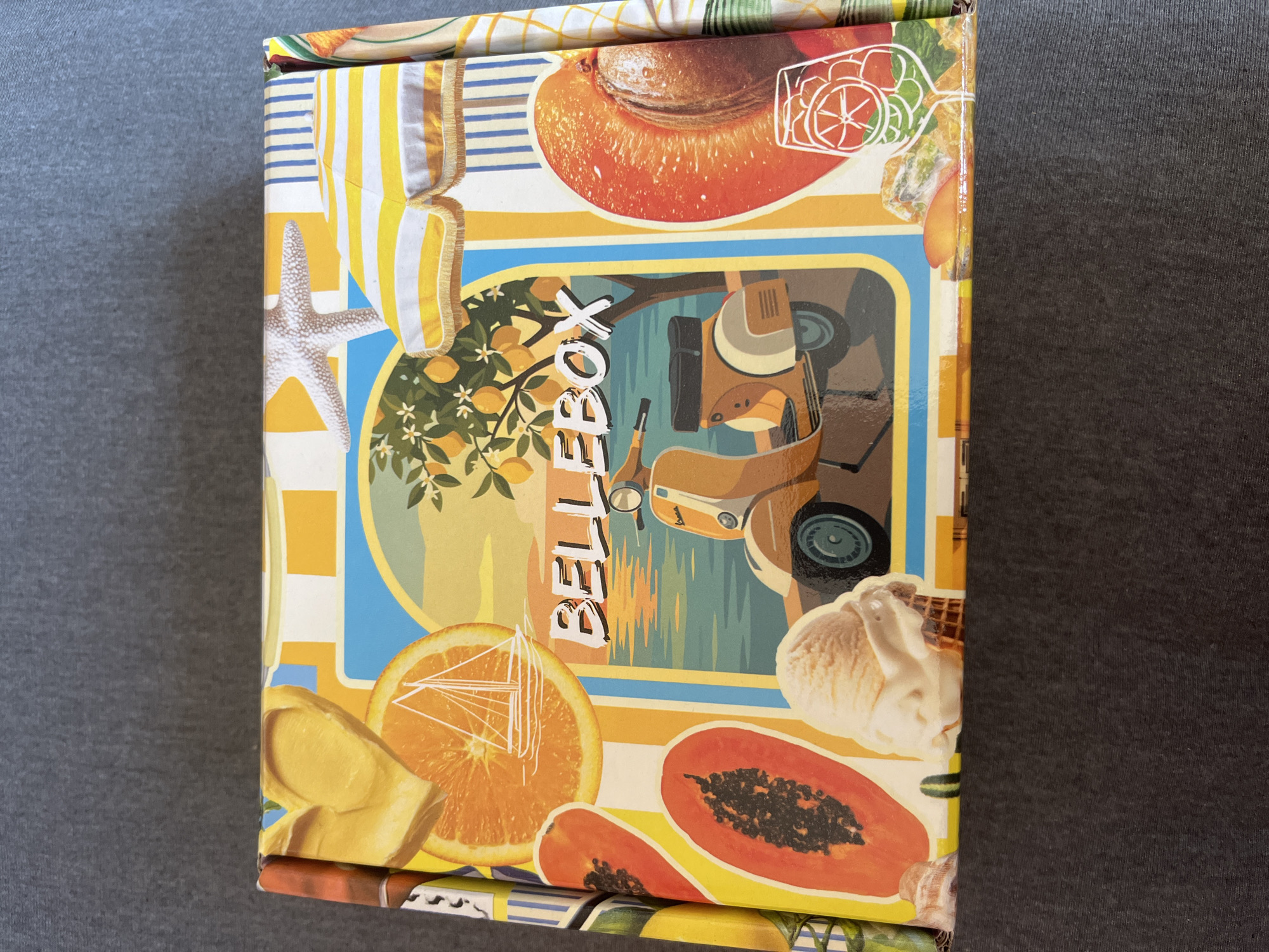 A rectangular box with a collage of fruit and summer-themed illustrations is displayed on a dark gray textured surface. The box features images of sliced oranges, papayas, an apricot, a glass of iced beverage, an ice cream cone, a yellow Vespa scooter, a sailboat, lemon trees, a starfish, and striped beach umbrellas. The text "BELLEBOX" is prominently displayed in white letters with a black outline on the front of the box. There are no people or animals visible in the image. The scene is indoors, with the box positioned flat on a surface that appears to be a tabletop or floor. The lighting suggests an overhead light source, creating soft shadows.