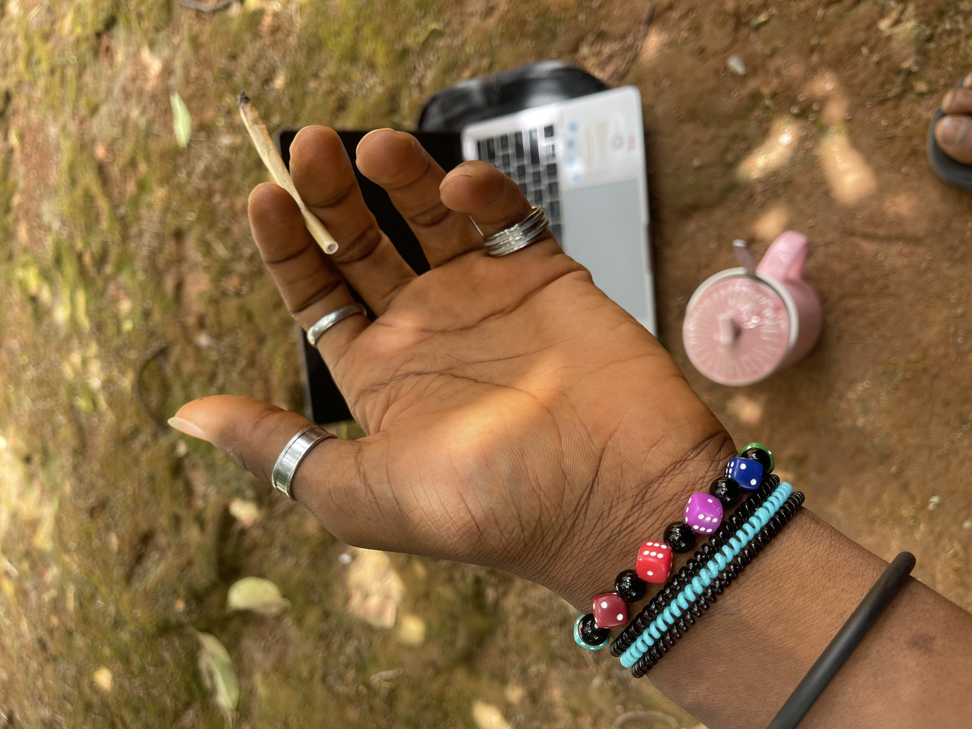 A dark-skinned person's hand holds a lit cigarette between the index and middle finger. The hand has silver rings on the thumb, index, and middle fingers. The wrist wears several bracelets, including one made of dice beads. A laptop, a pink smoothie blender, and part of a pair of sandals are visible on the ground nearby, likely outdoors on red soil. The setting appears to be an outdoor area in Jos, Nigeria.