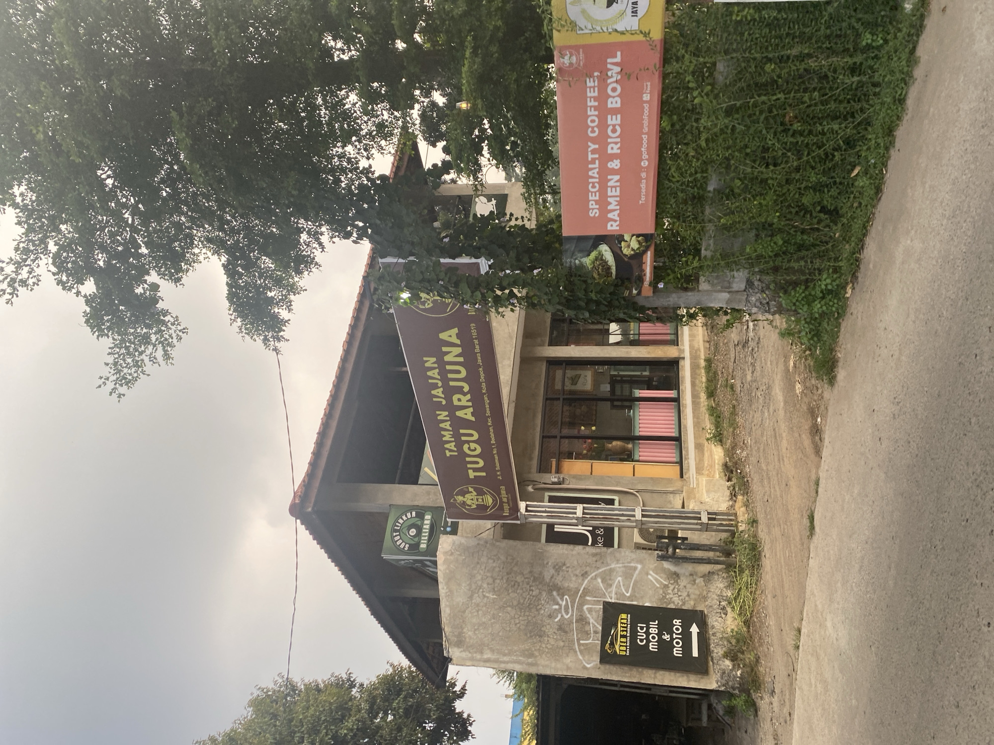 The image captures a commercial building in Depok, Indonesia, featuring a prominent sign for "TAMAN JAJAN TUGU ARJUNA." The building has a weathered, concrete exterior and a sloped, tiled roof. In front of the building, several signs advertise services. To the right, an orange banner reads "SPECIALTY COFFEE, RAMEN & RICE BOWL" and mentions availability on GoFood and GrabFood. To the left, a black sign with white text states "UBER STEAM CARS & BIKES WASHING STATION" and below it "CUCI MOBIL & MOTOR," indicating car and motorcycle washing services. A smaller green sign above the main entrance says "SOROT LINKUP BILLIARD." The entrance appears to be a glass-enclosed storefront with pink and white detailing. Lush green vegetation covers parts of the building's exterior and a wall to the right. A tree with dense foliage is visible in the upper right portion of the image. The ground in front of the building is a mix of dirt and paved road. The sky is overcast.