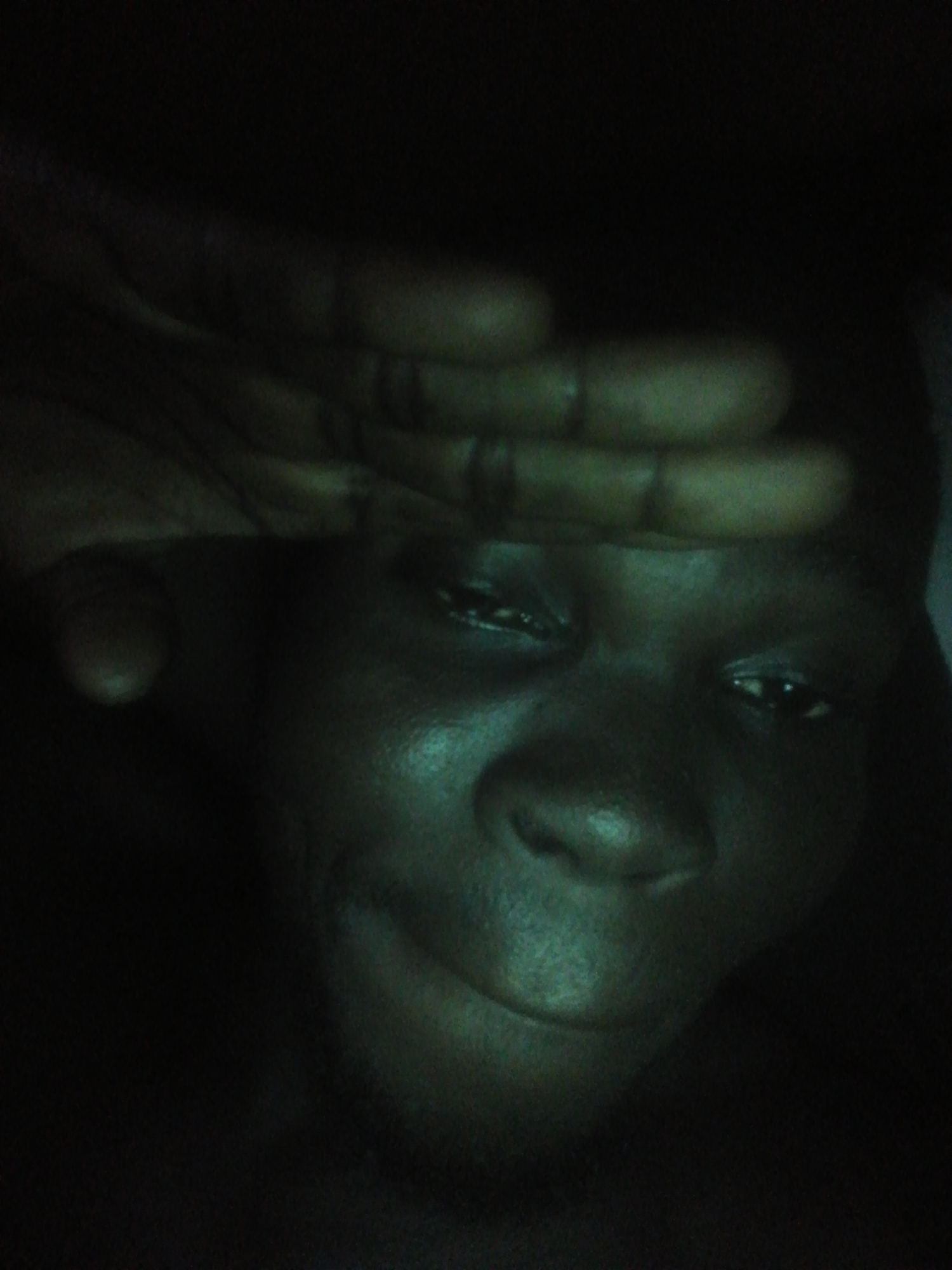 A close-up photo depicts a dark-skinned person with their hand raised to their forehead, possibly shielding their eyes. The lighting is poor, making it difficult to discern details. The background is black. The location is unknown.