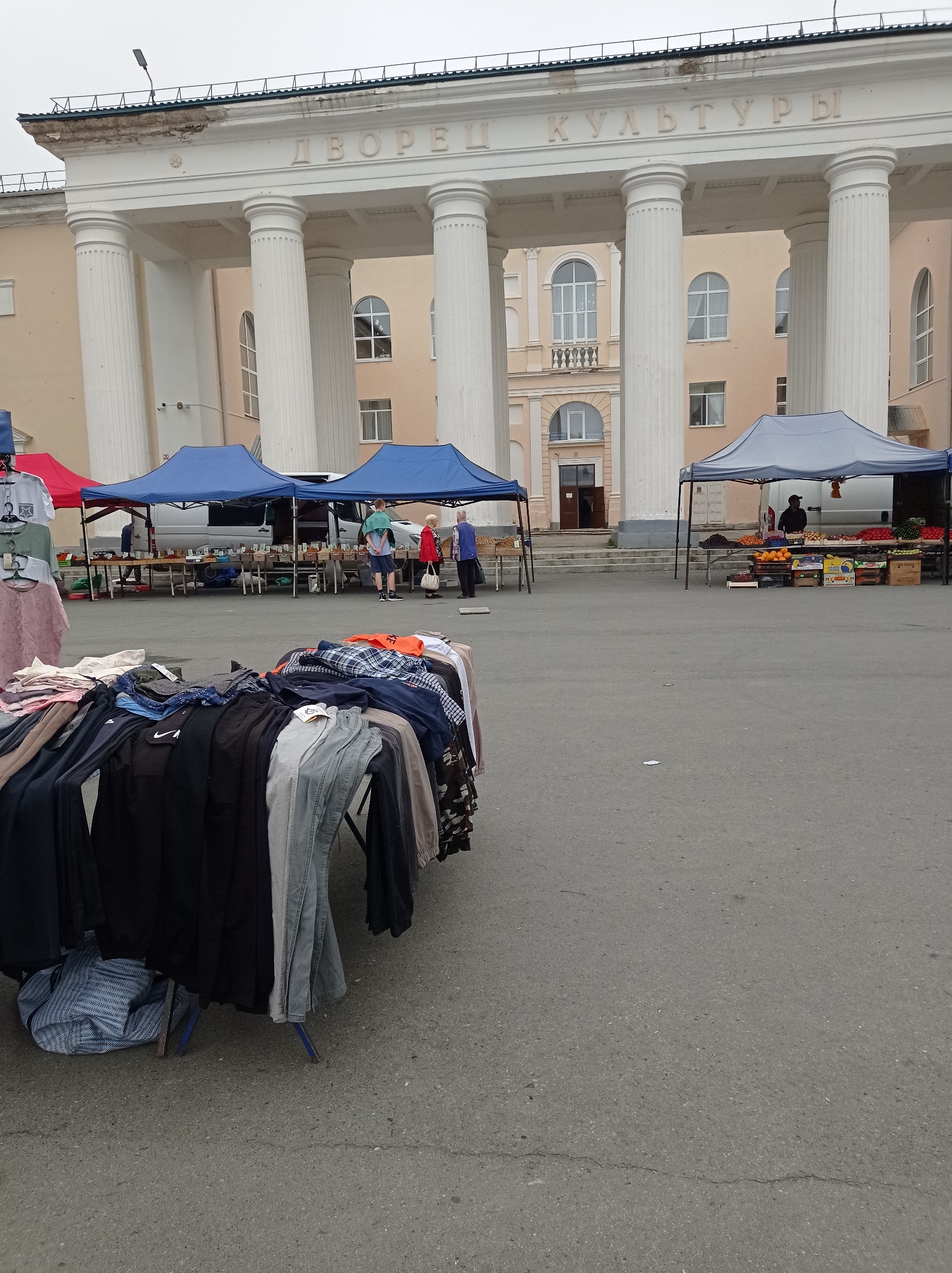This is an outdoor image of a market in front of a building with large columns and the Cyrillic inscription "ДВОРЕЦ КУЛЬТУРЫ" which translates to "Palace of Culture". The building appears to be constructed of light-colored stone or stucco. In the foreground, there are several tables displaying clothing for sale, including pants, shirts, and a checkered jacket. Behind the clothing stalls, there are two blue tents set up, under which vendors are selling goods. Further back, to the right, is a gray tent with a vendor selling fruits and vegetables, including oranges, grapes, and tomatoes. Several people are visible at the market: two individuals are standing near the blue tents, one wearing a green shirt and the other a red jacket. Another person is standing at the fruit stand. The ground is paved with asphalt. The overall impression is a bustling outdoor market scene in a city square.