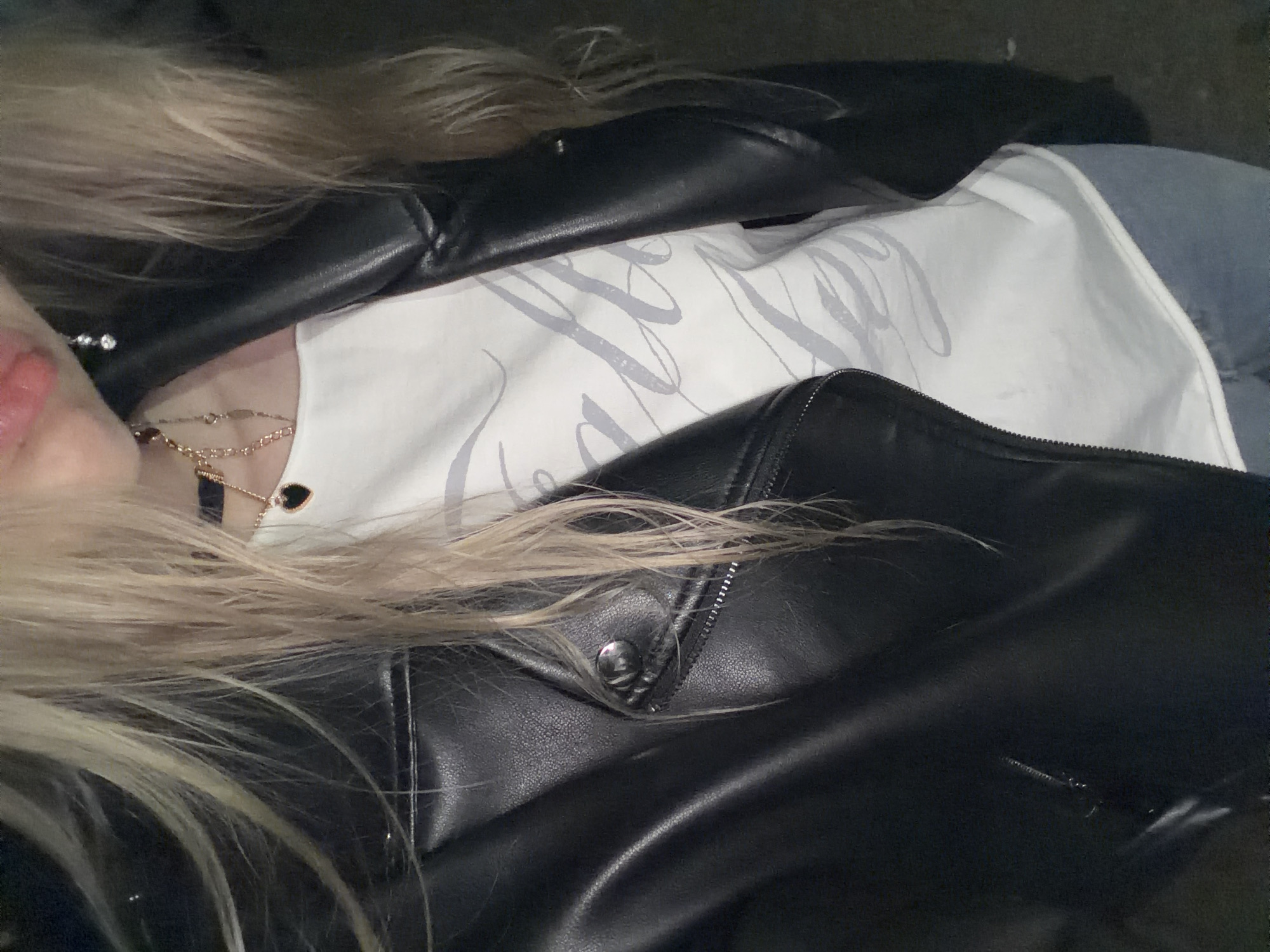 A close-up, chest-up view captures an individual with long, straight, light blonde hair and visible dark roots, worn loose over the shoulders. The individual's lips are slightly parted and adorned with pink lip gloss. They are wearing a black faux leather biker jacket with a silver zipper and a snap button detail on the lapel. Beneath the jacket, a white t-shirt is visible, featuring a light gray, stylized script graphic. A black choker necklace, a layered gold-toned necklace with a small black heart pendant, and a silver-toned dangle earring with clear stones are worn as accessories. The lower body is partially visible, showing light blue denim jeans. The background is dark and indistinct, suggesting low light conditions at an unspecified location in the city of Unknown, Unknown.