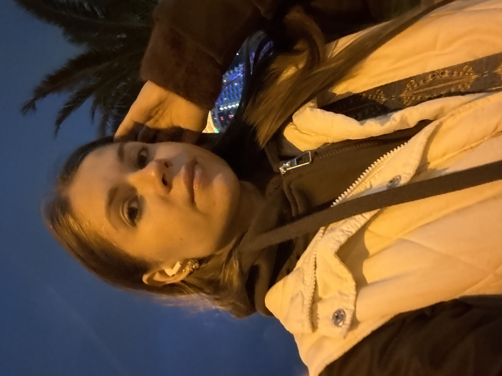 The image, taken at night or late dusk, features a young woman in a selfie-style shot, rotated 90 degrees counter-clockwise. She has long, dark hair visible on the left side of her face and is looking towards the upper part of the frame. Her right hand is raised, with fingers resting gently near her forehead. She is wearing a dark top, possibly a hoodie, layered under a light-colored, likely cream or beige, quilted jacket. The jacket has a distinct dark patterned strap or band visible across the chest area and a silver snap button near the collar. A small, light-colored object, possibly an earbud or earring, is visible in her right ear.
The background is a dark blue sky, indicating the time of day. To the upper left (original orientation), the dark silhouette of palm fronds suggests a warm climate. To the upper right, blurred, colorful city lights, predominantly blues and reds, are visible, hinting at an urban setting. This aligns with the provided location context of Batumi, Georgia, a coastal city known for its palm trees and illuminated architecture. There is no visible text. The overall mood appears calm.