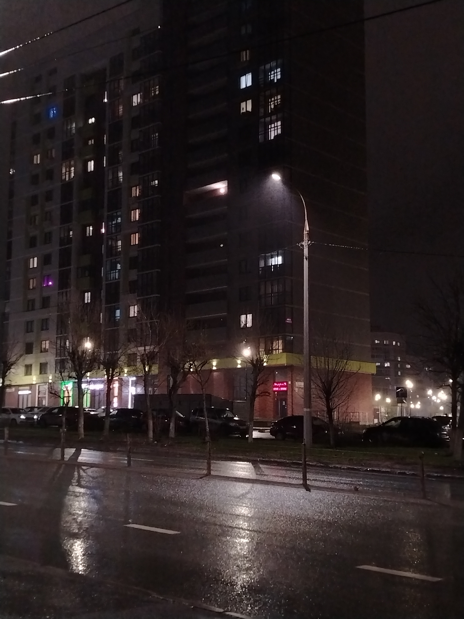 The image shows a nighttime street scene in Izhevsk, Russia. A multi-story apartment building with numerous illuminated windows dominates the left and center of the frame. A street lamp illuminates a wet road with painted white lines. Several cars are parked along the curb. Small trees line the street. It appears to be raining.