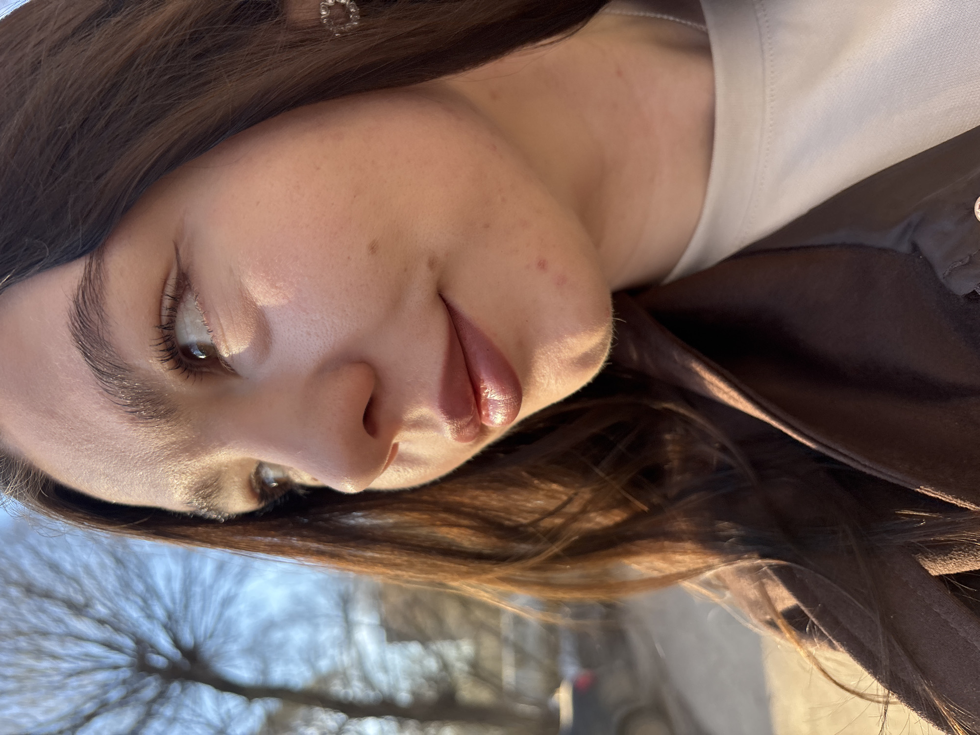 The image presents a close-up, diagonally oriented portrait of a young individual, likely female, in an outdoor setting during the daytime. Her head is tilted, and she is looking to the left of the frame. Her complexion is fair, with natural skin texture, including some freckles and minor blemishes visible on her cheek and nose area. She has distinct, dark eyebrows and long, dark eyelashes framing her visible eye, which appears to be light brown or amber in color. Her lips are slightly parted, showing a soft, possibly glossy, pinkish-brown lip color.
She wears a light-colored, possibly white, top beneath a dark brown or black jacket, hints of which are visible in the lower right portion of the image. Her hair is long, dark brown, and appears to be softly styled around her face. A small, circular, sparkling earring is visible in her right ear, peeking through her hair.
The background is highly blurred, indicating a shallow depth of field. It depicts out-of-focus tree branches against a light blue and grey sky, suggesting a park, street, or other natural outdoor environment. The bright, natural light creates subtle highlights on her skin and hair, consistent with sunny or partly cloudy weather. The image captures a casual, spontaneous moment. No visible text is present in the image. The general location is Perm, Russia.
