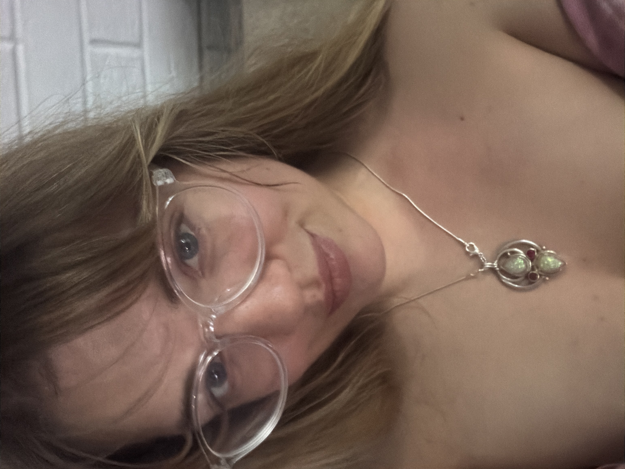 A woman with blonde hair is wearing glasses and a necklace. The location is Большая Шильна, Russia. The environment appears to be an indoor setting with a white brick wall in the background.