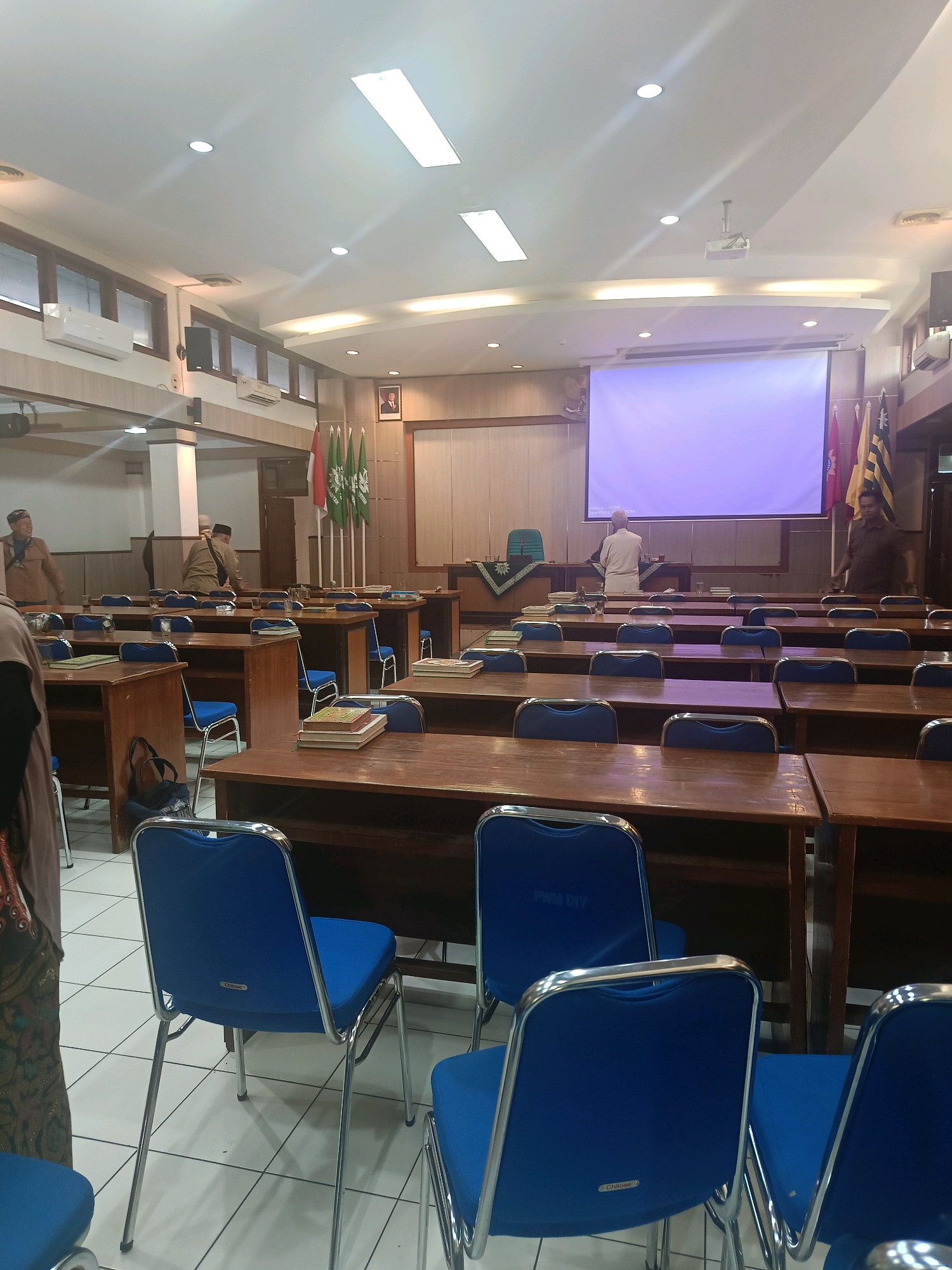 The image depicts an indoor view of a classroom or lecture hall, likely within a university or educational institution in Yogyakarta, Indonesia. The room is furnished with rows of wooden desks and blue-seated chairs. In the foreground, several blue chairs with chrome frames are visible, some with the "Chitose" brand label on the backrest. Stacked books are present on some of the desks.
At the front of the room, a large projection screen displays a blank purple image. A podium stands to the left of the screen. A man in a white shirt is visible behind the podium, facing away from the camera. Several flags, including a red and white Indonesian flag and green flags with symbols, are displayed behind the podium. Two men in similar brown uniforms and caps are standing near the entrance on the left side of the room, and another man in a dark shirt is walking away from the camera on the right side, near the projection screen.
The environment appears to be well-lit by fluorescent ceiling lights and natural light coming from windows. Air conditioning units are visible mounted on the wall above the windows. The floor is tiled with a white checkered pattern. The overall impression is that of a functional educational space prepared for a lecture or meeting.
