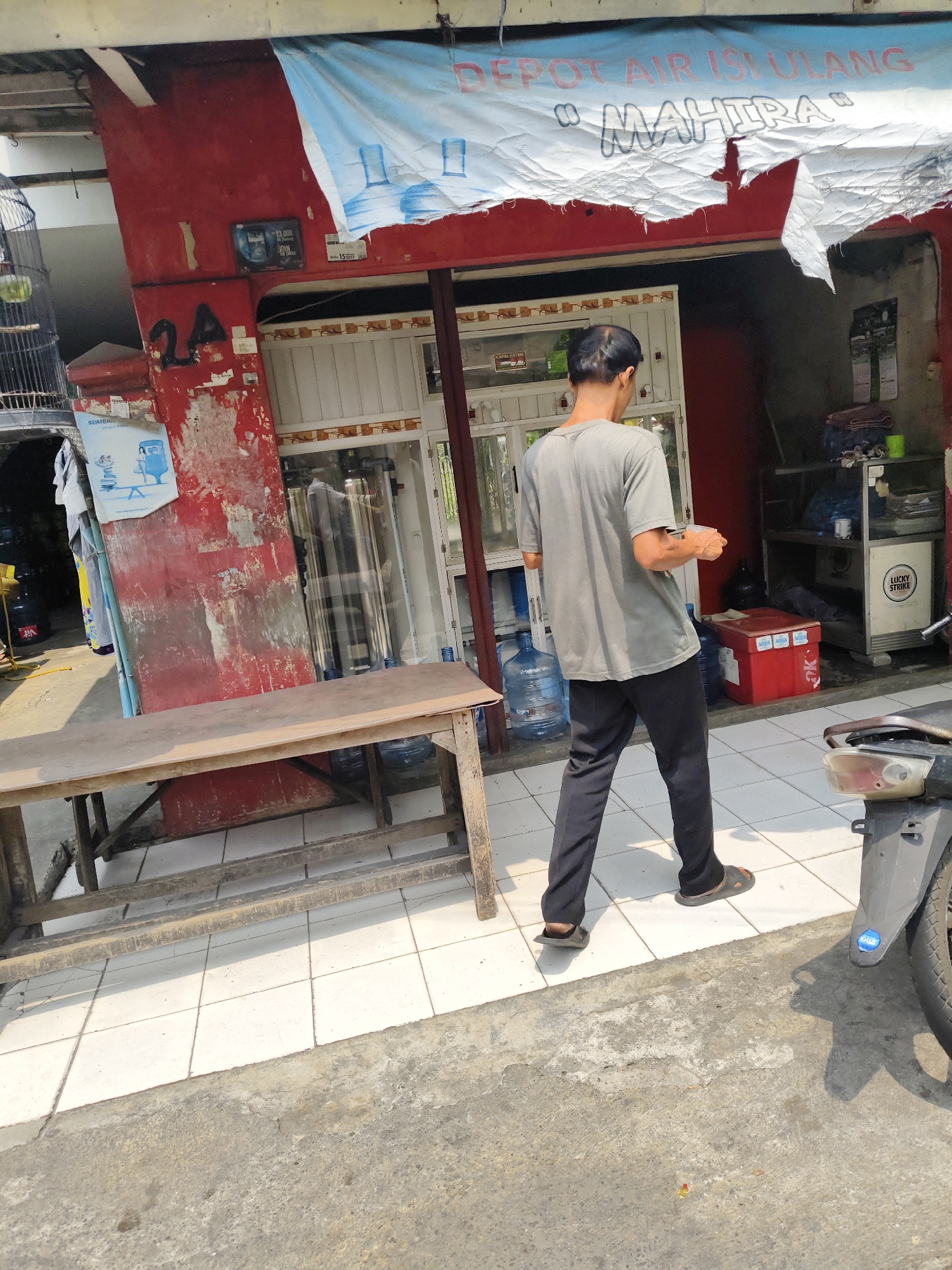 The image depicts a street scene in South Jakarta, Indonesia, likely outside a water refill station. A man, appearing to be in his 30s or 40s, with short dark hair and wearing a grey t-shirt and black pants, is walking away from the camera. He is holding a clear plastic cup in his right hand. In the background, a red building with peeling paint forms the facade of a shop. A large blue and white banner hangs above the entrance, with text that appears to read "DEPOT AIR ISI ULANG MAHITRA" which translates to "MAHITRA WATER REFILL STATION". A birdcage is visible to the left of the entrance. In front of the shop, a wooden table and benches are set up, and several large water bottles are stacked. Further to the right, a motorcycle is parked. The ground is paved with white tiles and concrete. The overall impression is of a typical urban commercial area in Indonesia.