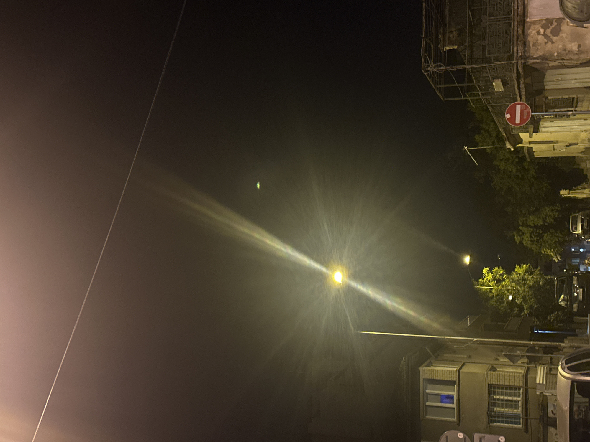 This is a nighttime photograph taken in Varna, Bulgaria. The scene captures a street with buildings, trees, and vehicles illuminated by streetlights. A prominent street lamp in the center of the image casts a bright, starburst-like glare, with a thin overhead wire visible above it. To the left, a multi-story building with windows of varying designs is partially visible. On the right side of the street, another building with a weathered facade and a metal structure above it stands. A red and white "no entry" traffic sign is mounted on a pole in front of this building. Several cars are parked along the street, including a black van and a white vehicle in the foreground. Trees are present on both sides of the street, some with yellow lights emanating from them, possibly from street lamps or decorative lighting. The sky is dark, with a faint, small, possibly distant, green light visible. The overall impression is of a residential street at night, with the characteristic urban elements of Varna.