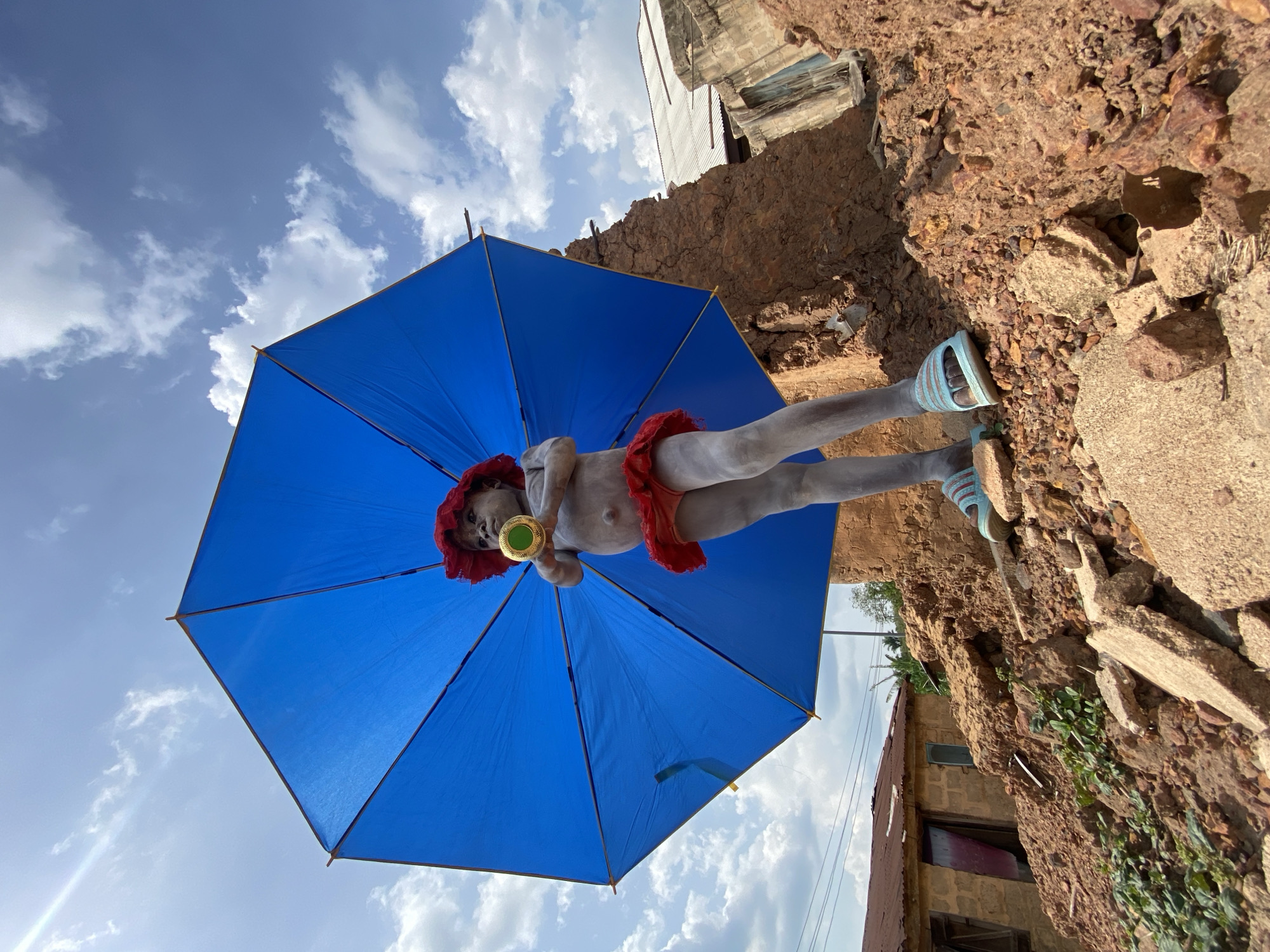A figure stands on a brown, rocky, and somewhat dilapidated surface in Igbara, Nigeria. The person, painted white, wears a red headdress and a red frill around the waist. They are holding a green and gold object, possibly a musical instrument, to their mouth. The figure is positioned beneath a bright blue umbrella. The background includes a cloudy blue sky and parts of a building with a window. The surface the figure is standing on appears to be a weathered and broken urban environment.