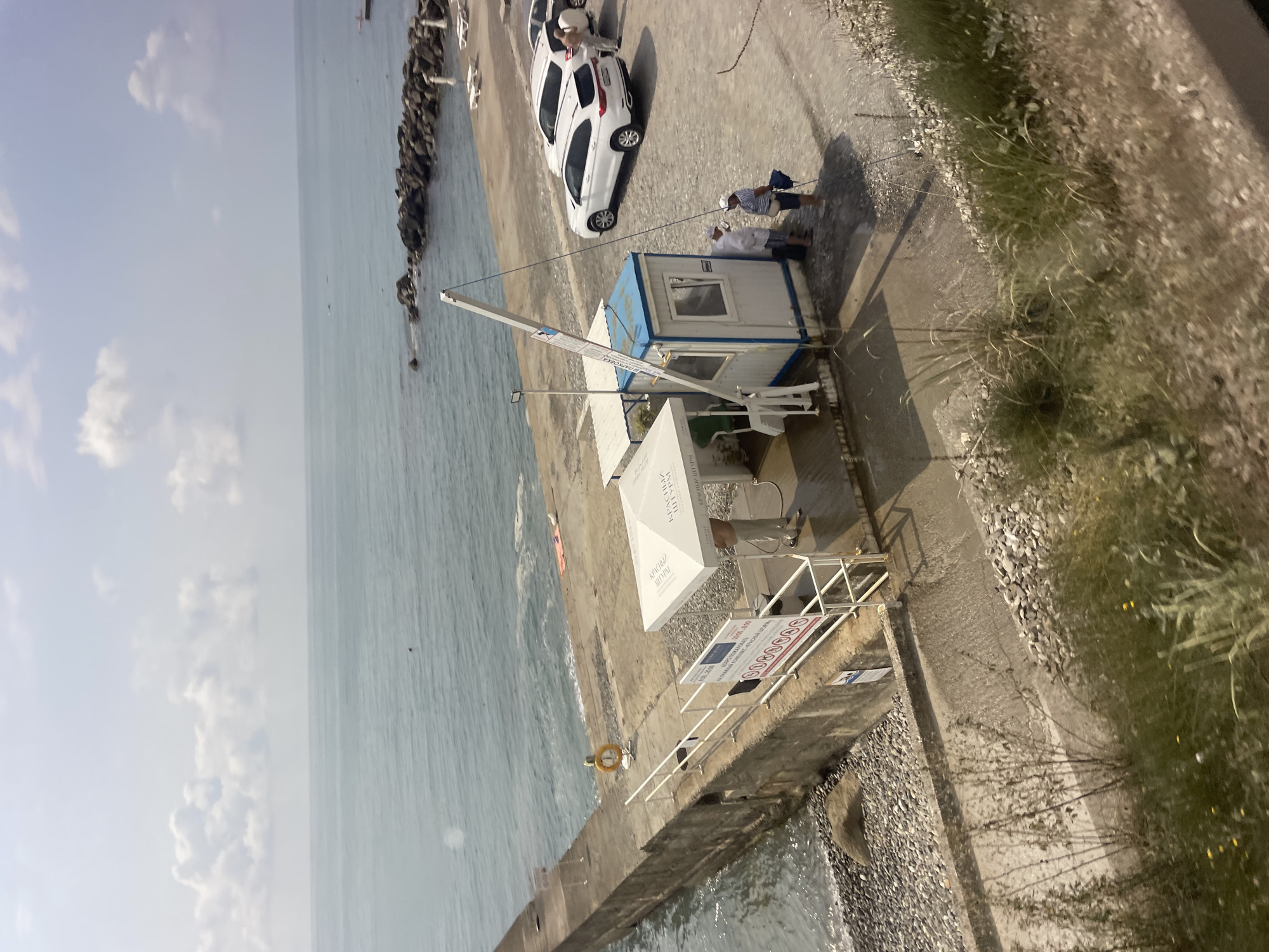 The image depicts a pier or seawall in Sochi, Russia, likely part of a recreational area or port. On the pier, a small blue and white booth with a barrier arm is situated, suggesting a controlled entry or access point. Two individuals, dressed in casual attire and hats, are seen standing near the booth, possibly interacting with each other or an attendant. Several white vehicles are parked on the paved area adjacent to the pier. In the background, the sea stretches out under a cloudy sky, with some rocks and a small jetty visible in the distance. The pier itself is constructed of concrete and lined with a railing and a life preserver ring. The area around the pier is covered in pebbles, typical of the Black Sea coast. The overall scene suggests a sunny day at a coastal location.
