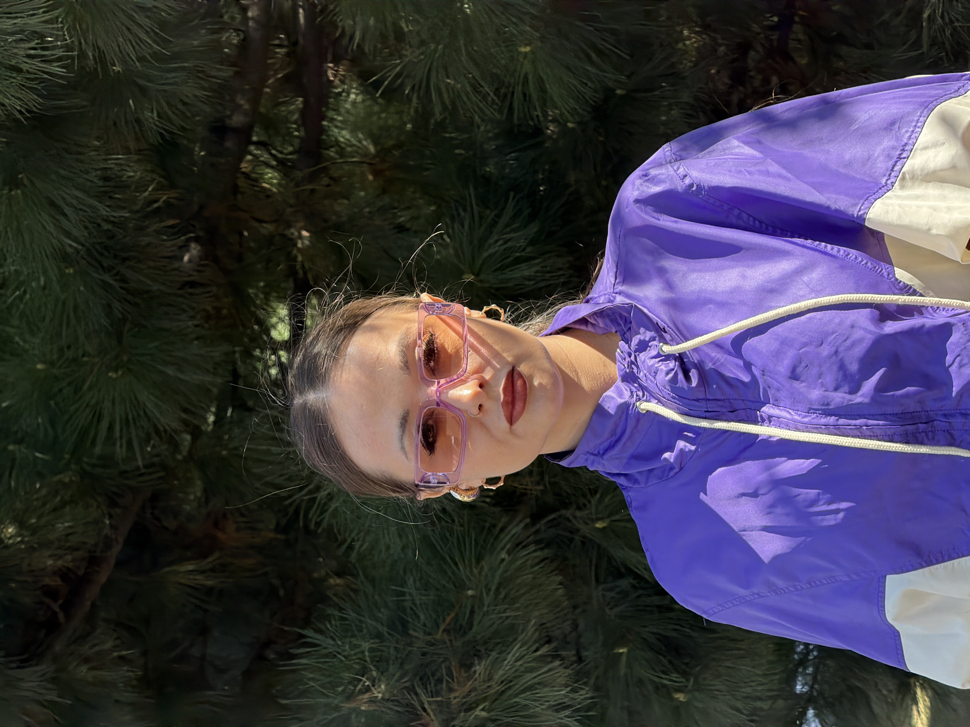 A young woman with her hair pulled back is standing in front of a coniferous tree. She is wearing pink sunglasses and a purple jacket with white trim. The setting appears to be outdoors, possibly in a park or forested area. The specific location within the city of Krasnoyarsk, Russia, cannot be determined from the image.