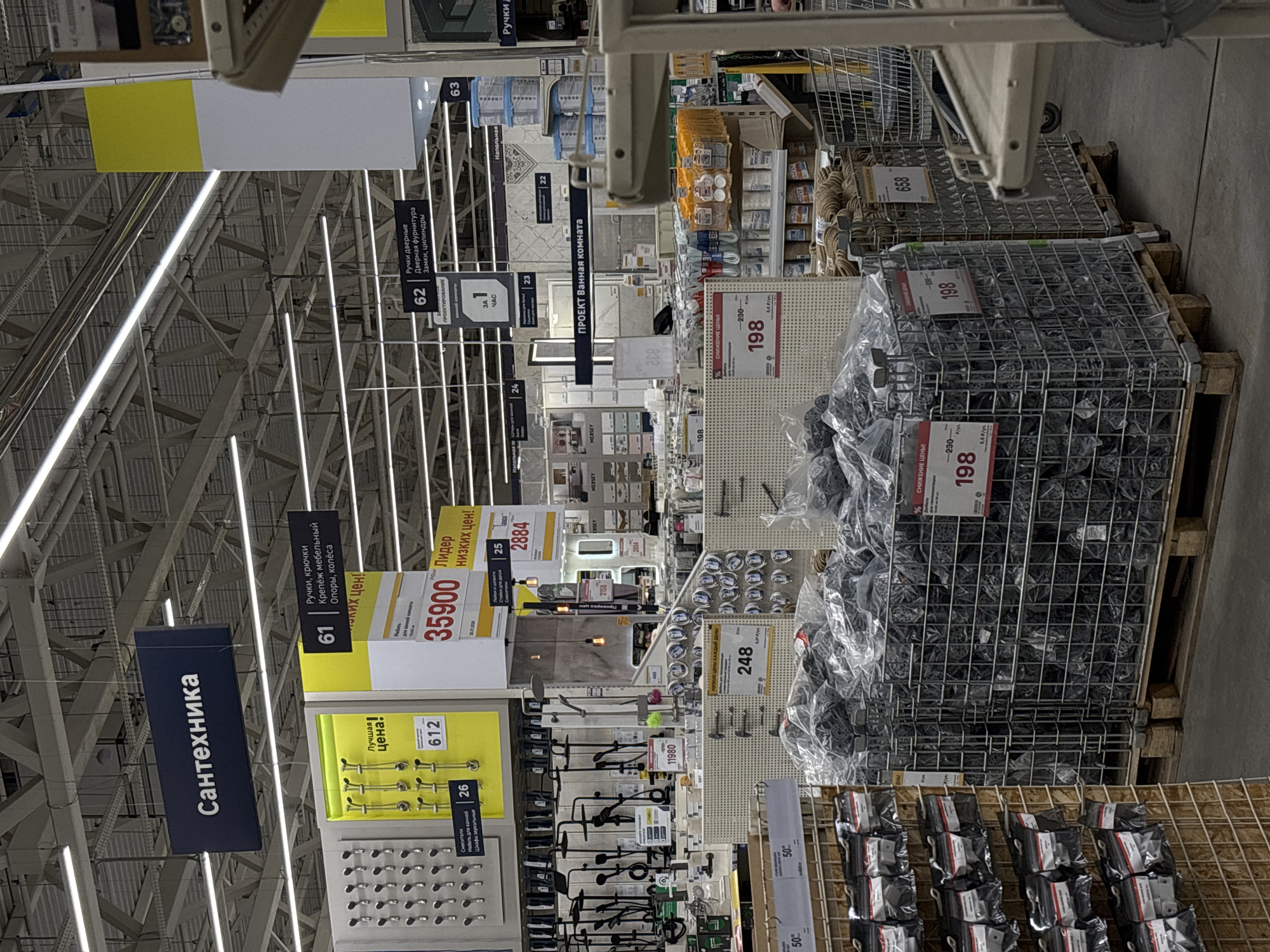 The image depicts the interior of a hardware store in Perm, Russia, likely a Leroy Merlin, as indicated by product pricing signs and product selection. The view is down an aisle, revealing rows of metal racks holding various hardware items. In the foreground, there is a large selection of furniture hardware such as knobs, pulls, and wheels, presented in wire mesh containers on pallets. Behind these, there are displays of bathroom fixtures, including faucets, showerheads, and mirrors. Signage is visible overhead, directing customers to different product categories like "Сантехника" (Sanitary Engineering), "Ручки дверные" (Door Handles), and "Проект ванная комната" (Bathroom Project). Price tags are prominent, indicating the cost of specific items, with prices such as 35900 and 198 displayed in red and black. The store is well-lit, with fluorescent lights illuminating the merchandise. The composition is slightly tilted.
