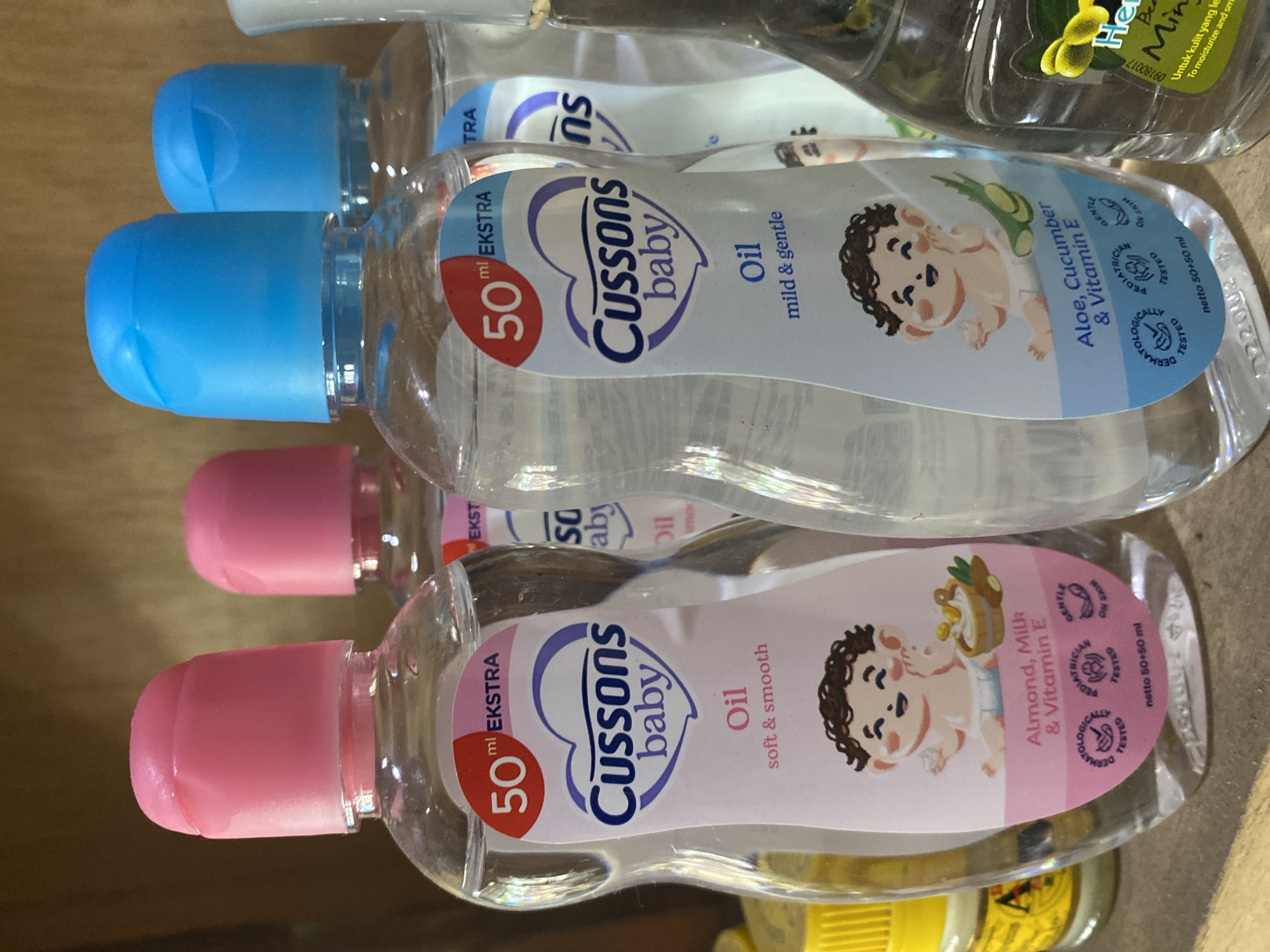 The image shows a close-up of several bottles of Cussons baby oil on a shelf. The bottles are clear and contain a light-colored liquid. The one in the foreground on the left has a pink cap and a label with a pink background. It says "50 ml EXTRA Cussons baby Oil soft & smooth" and features a cartoon drawing of a baby with the text "Almond, Milk & Vitamin E". The bottle in the foreground on the right has a blue cap and a label with a blue background. It says "50 ml EXTRA Cussons baby Oil mild & gentle" and features a similar baby drawing with the text "Aloe, Cucumber & Vitamin E". The bottles are arranged in a staggered manner, with portions of other bottles visible behind them. The background appears to be a wooden shelf. The lighting suggests an indoor environment. There are no people visible in the image. The location is not specified within the image itself, and cannot be determined from the visual information provided.