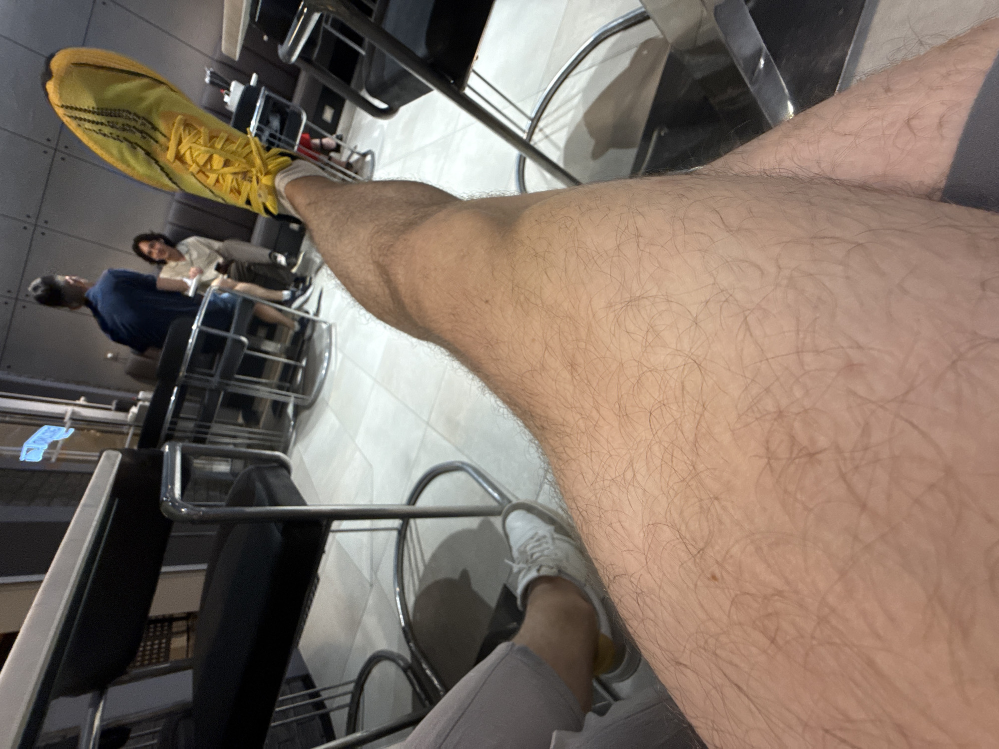 The photograph, taken from an elevated perspective, appears to be an amateur shot captured in an interior setting, possibly a cafe or restaurant. The primary focus is on a person's leg, positioned in the foreground, extending towards the camera. The leg is hairy, suggesting it belongs to an adult male. The person is wearing grey trousers and white shoes with a gold-colored accent.
In the background, blurred figures of two people are visible seated at a table. A man in a blue shirt is slightly visible. A woman in a light-colored shirt sits next to the man.
The environment suggests an urban location, as the photograph was taken in Tashkent, Uzbekistan. The furniture is modern, featuring metal barstools and tables. The floor is tiled. The lighting is artificial, and the overall impression is of a casual, public space.