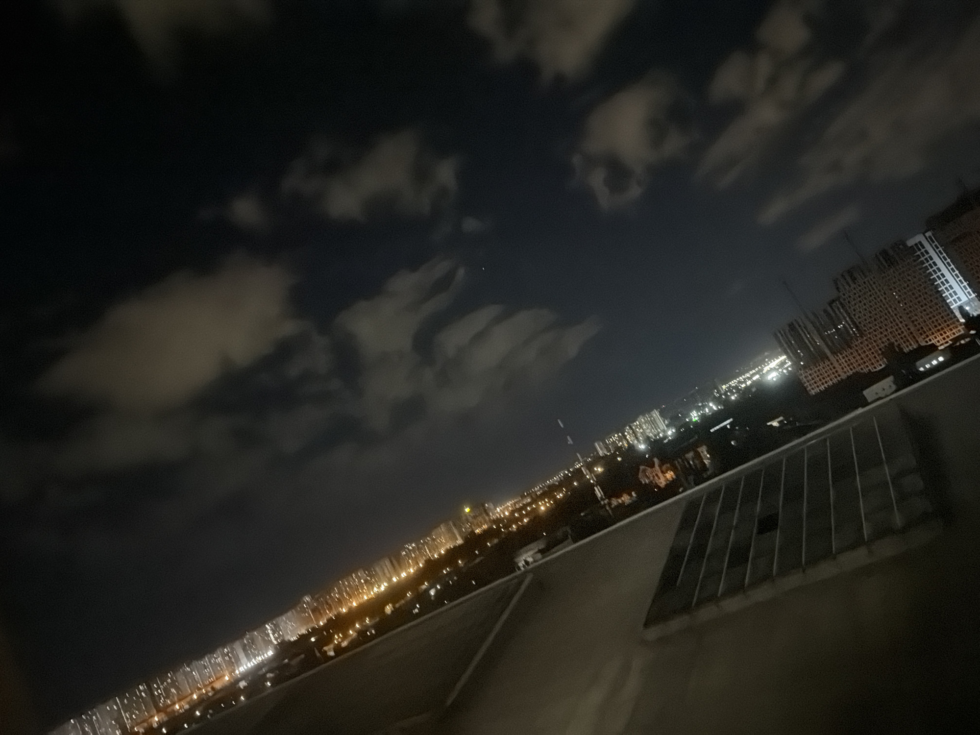 This is a nighttime aerial view of a city, possibly Kyiv, Ukraine, taken from a high vantage point. The cityscape is illuminated by a vast expanse of city lights, stretching into the distance. The buildings are densely packed, with numerous windows reflecting the light. The sky is dark with scattered clouds that are faintly illuminated by the city glow. A large ventilation grate is visible in the foreground, attached to a wall. The perspective is tilted, giving a dramatic and somewhat disorienting view of the urban environment. No people or specific actions are discernible in this image.